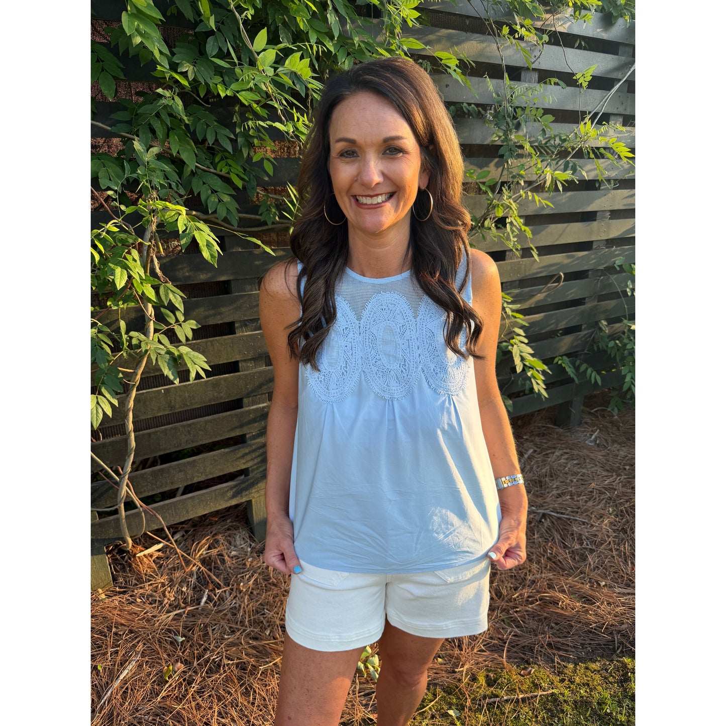Woman wearing a light blue sleeveless top and white shorts standing in front of a wooden fence with greenery.
