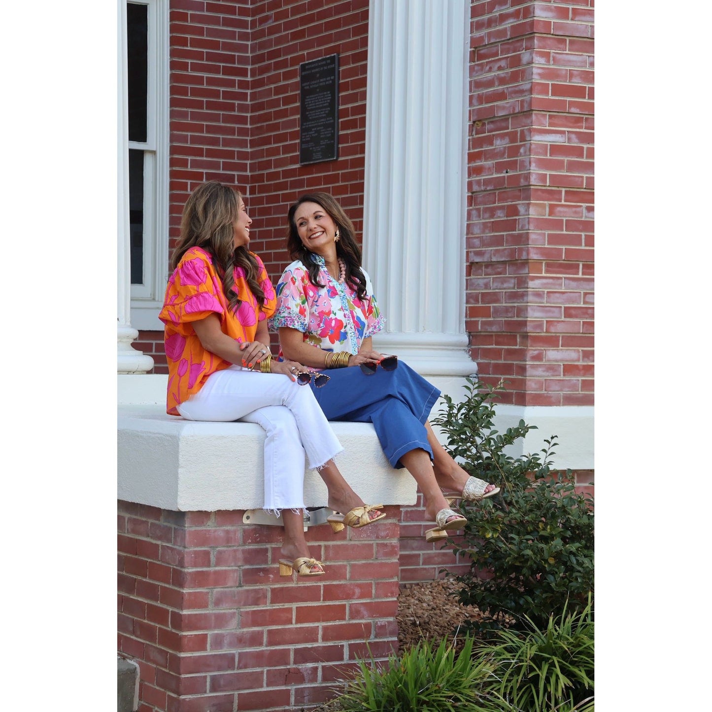 Two women sitting on a brick building's steps, smiling and enjoying each other's company.