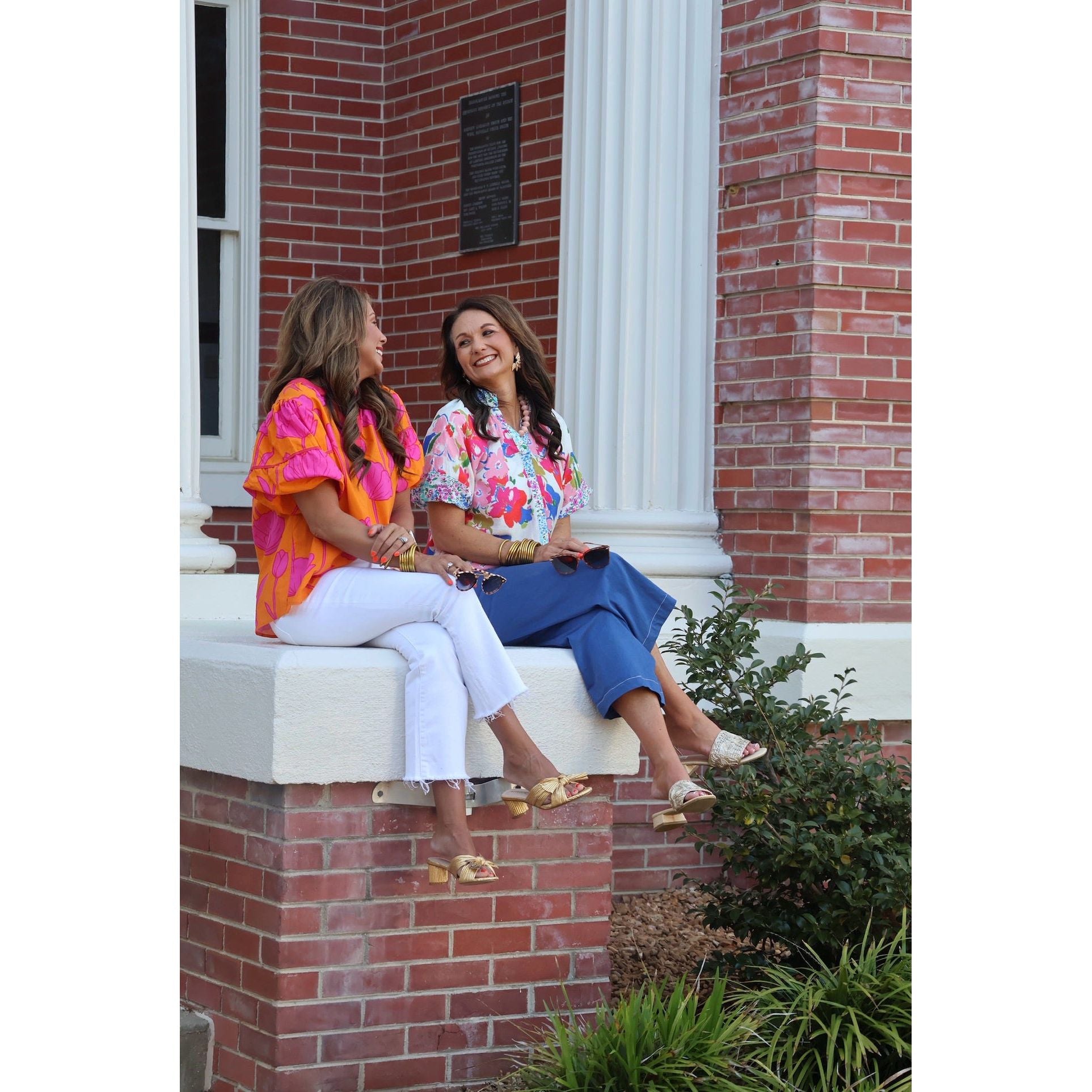 Two women sitting on a brick building's steps, smiling and enjoying each other's company.