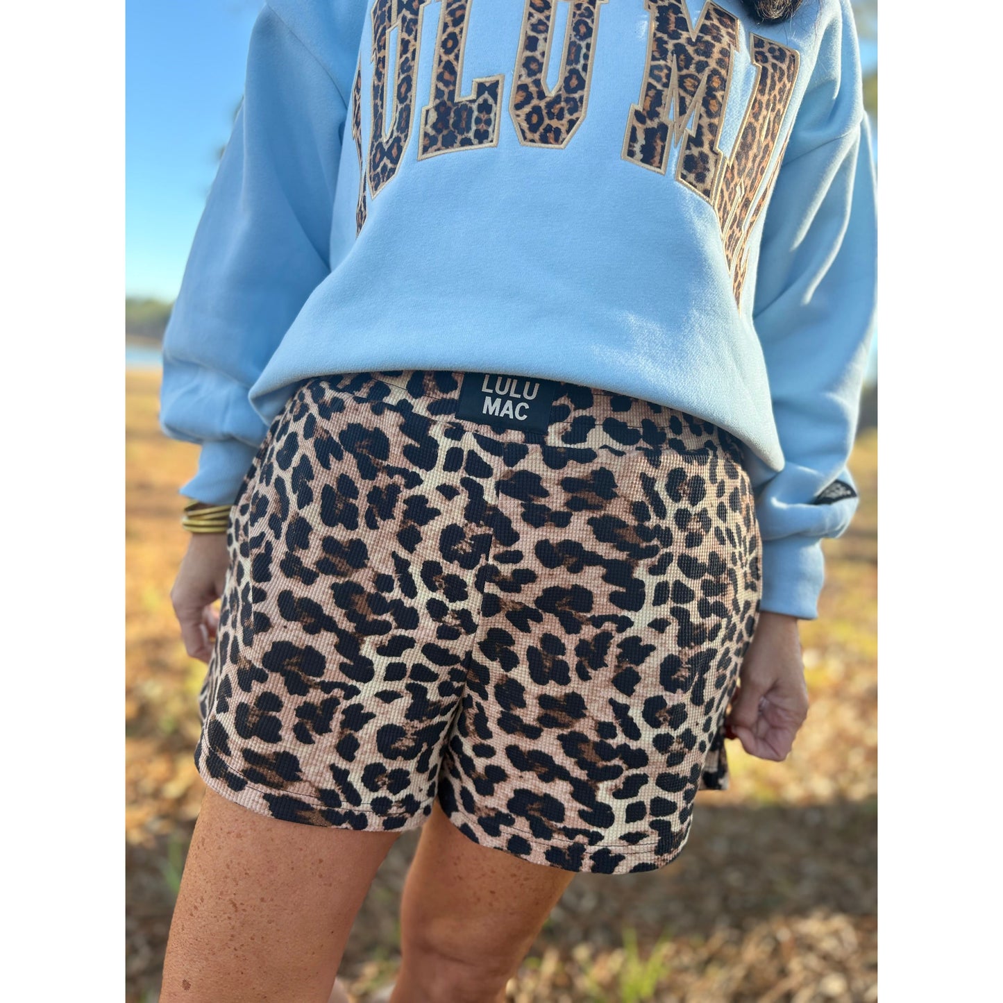 Person wearing a light blue sweatshirt with leopard print text and leopard print shorts, standing outdoors.