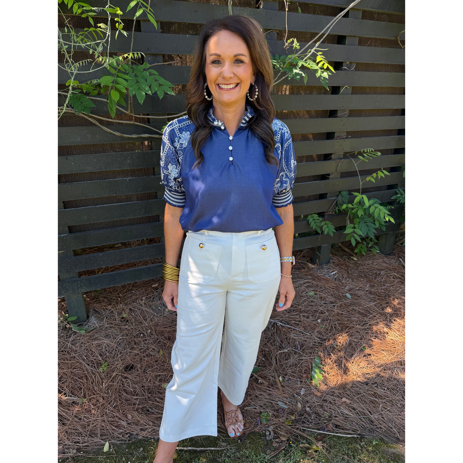 Woman wearing a blue top and white pants standing in front of a wooden fence with plants.