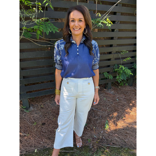 Woman wearing a blue top and white pants standing in front of a wooden fence with plants.