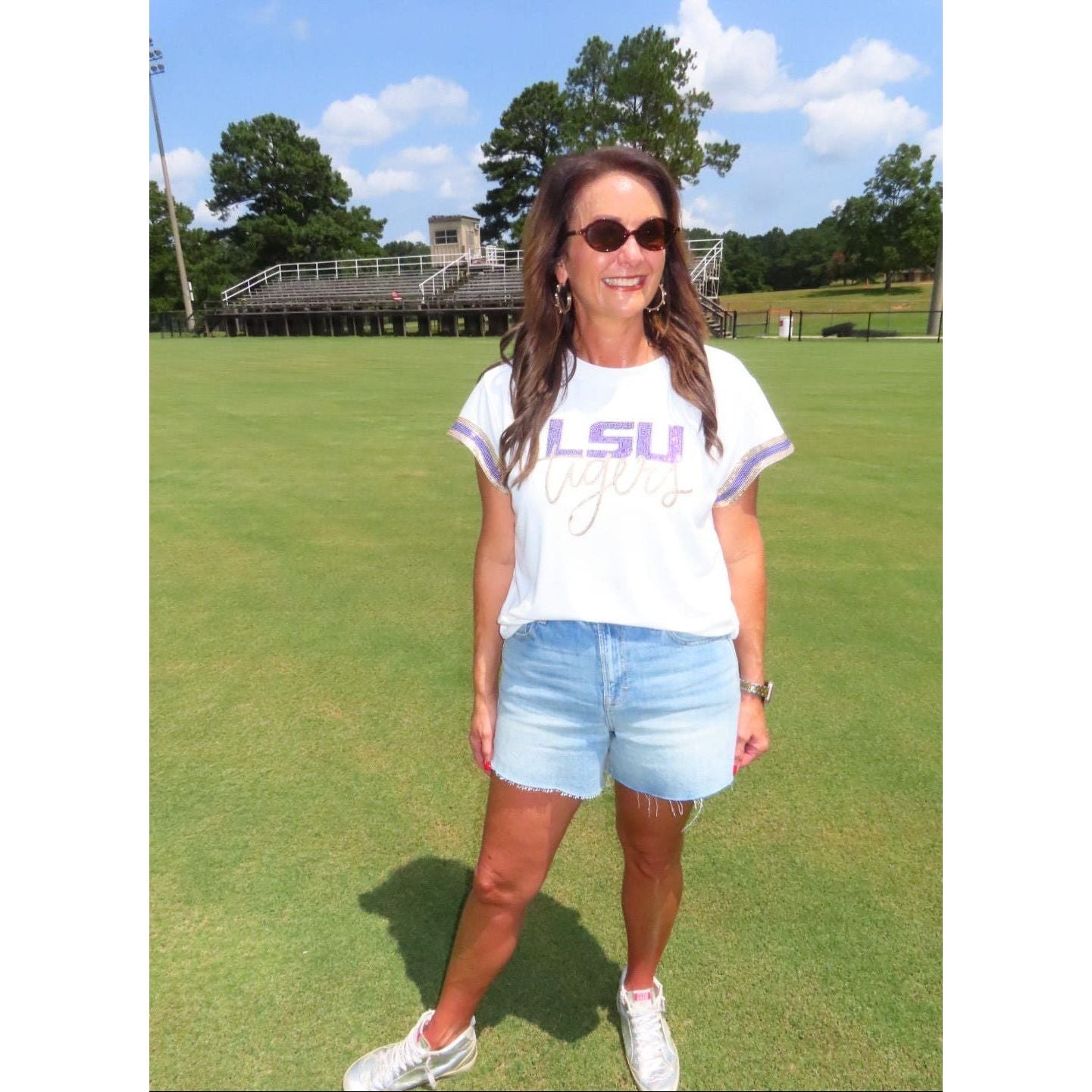 A woman standing on a green grass field wearing a white LSU Tigers graphic t-shirt, light blue denim shorts, and white sneakers.