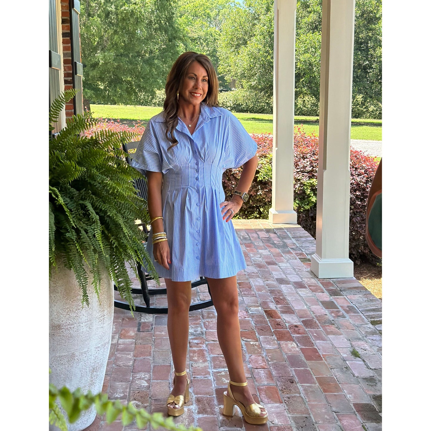 Woman in a light blue dress standing on a porch with greenery and a brick wall in the background.