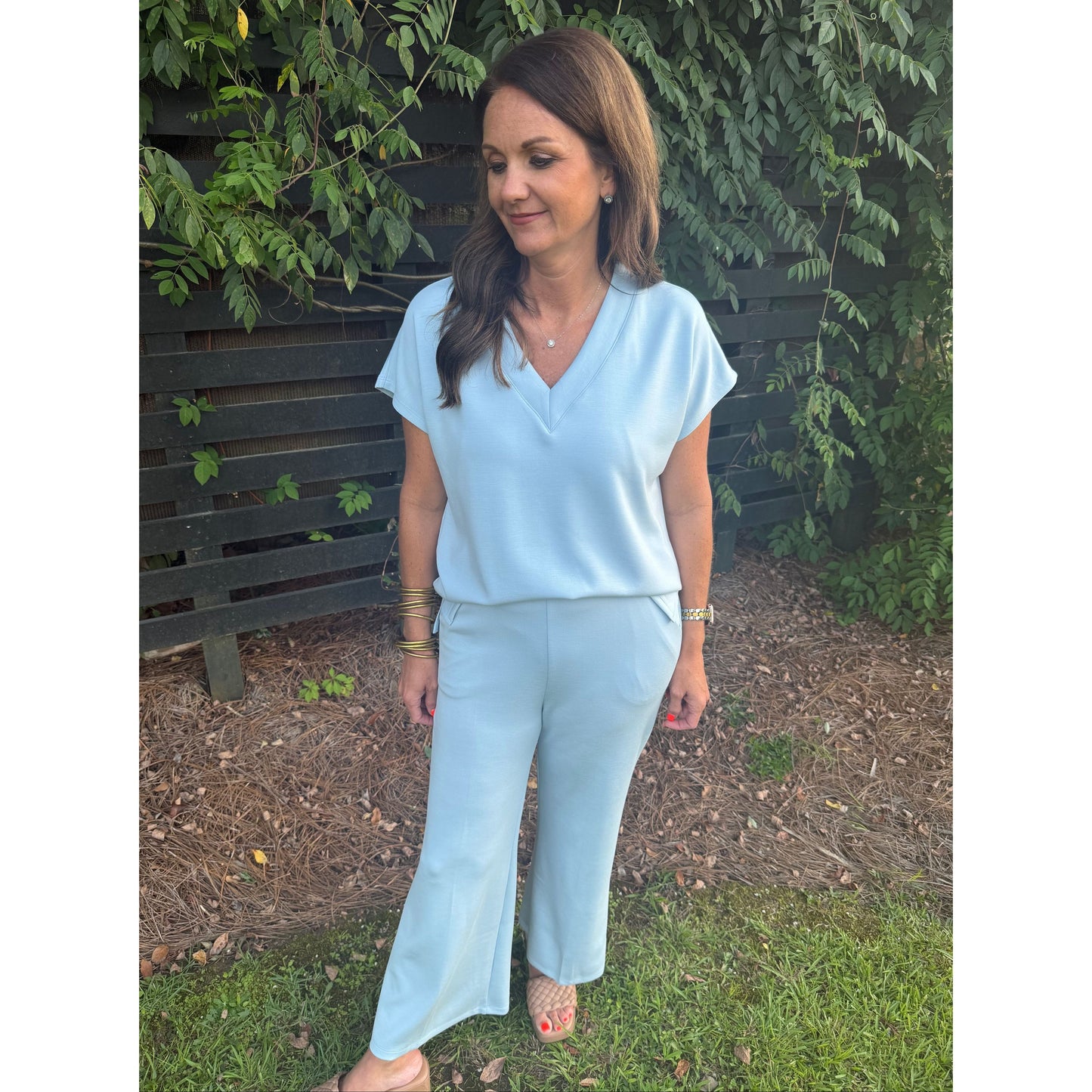 A woman wearing a light blue matching two-piece outfit consisting of a short-sleeve V-neck top and wide-leg pants, standing outdoors on a wooden bench with greenery and mulch in the background.