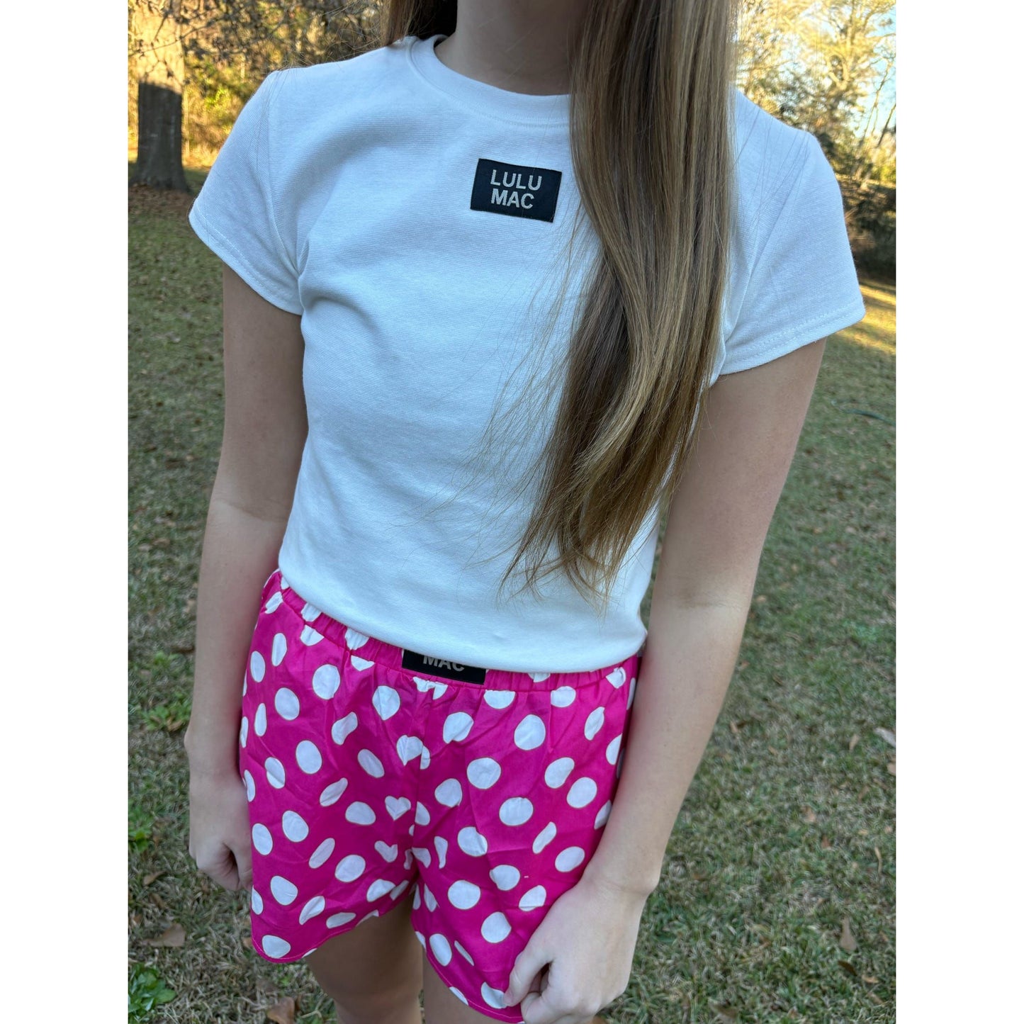 Person wearing a white t-shirt with 'LULU MAC' branding and pink polka dot shorts outdoors.
