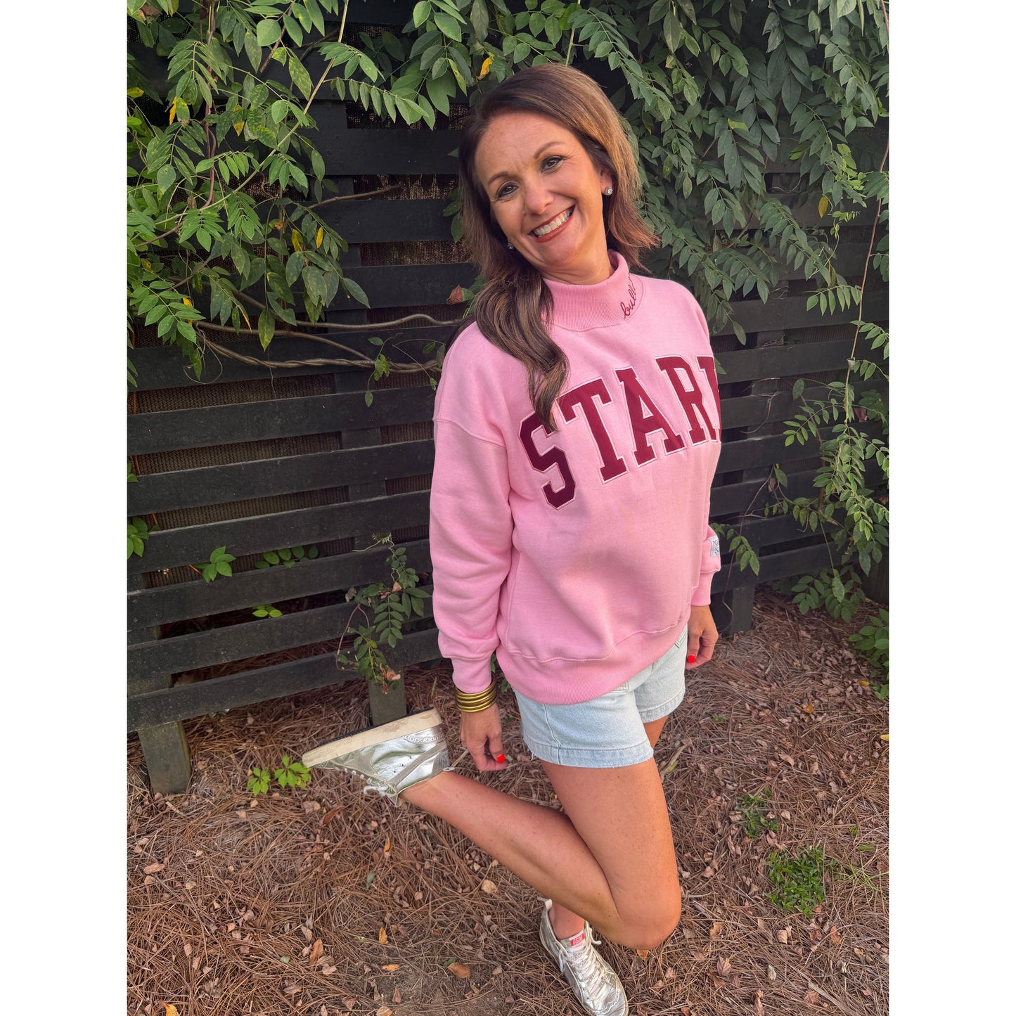 A woman posing outdoors in front of a dark wooden fence and green ivy, wearing a pink sweatshirt with 'START' printed in maroon varsity letters across the chest, light denim shorts, and silver sneakers.