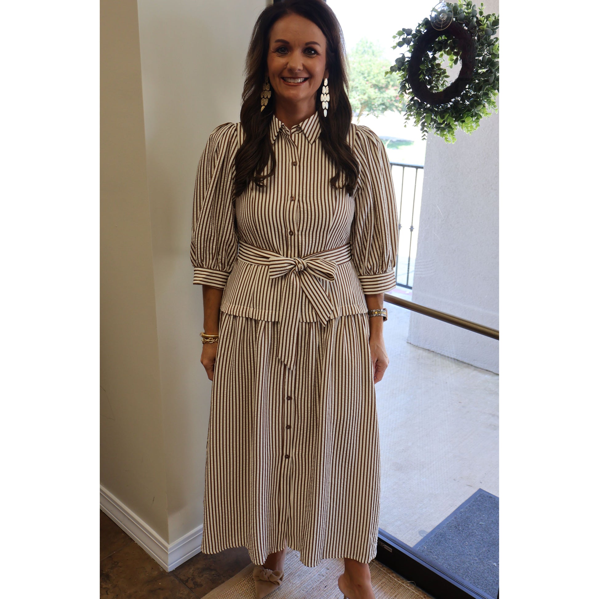A woman wearing a beige and brown vertically striped midi dress with puff sleeves, a button-down front, and a matching self-tie belt at the waist, standing in an indoor setting.