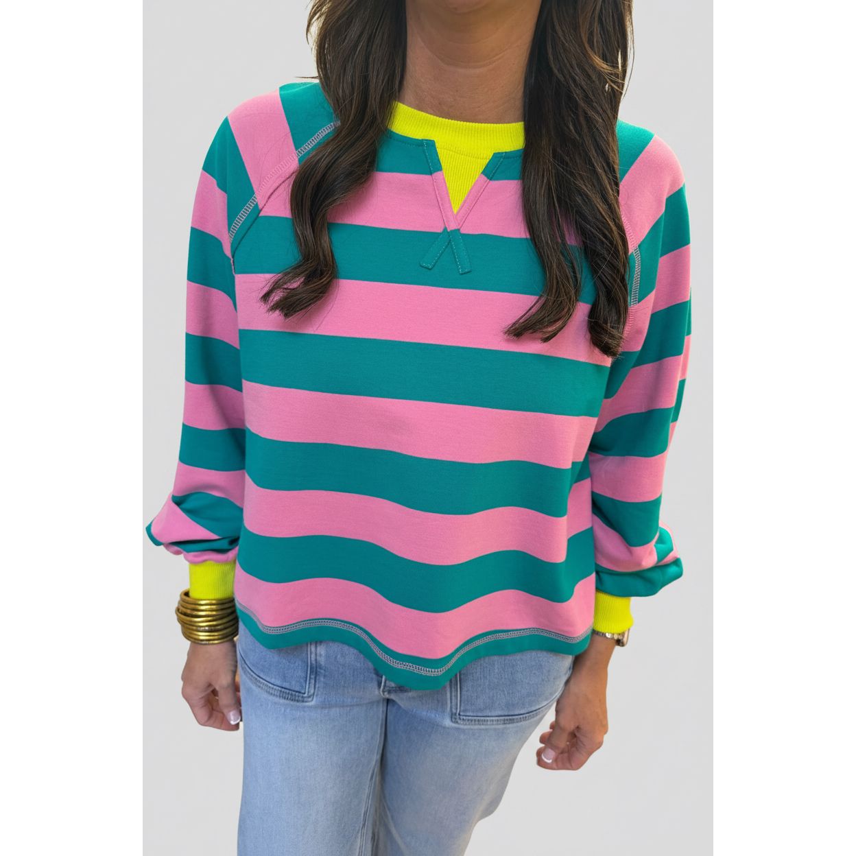 A woman wearing a pink and teal wavy striped long-sleeve top with a neon yellow V-neck collar, paired with light blue jeans.