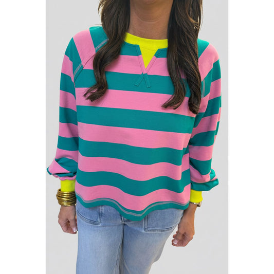 A woman wearing a pink and teal wavy striped long-sleeve top with a neon yellow V-neck collar, paired with light blue jeans.