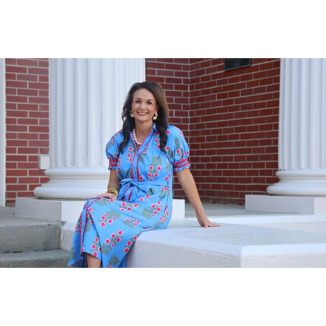 Woman in a blue floral dress sitting on steps with a brick building in the background