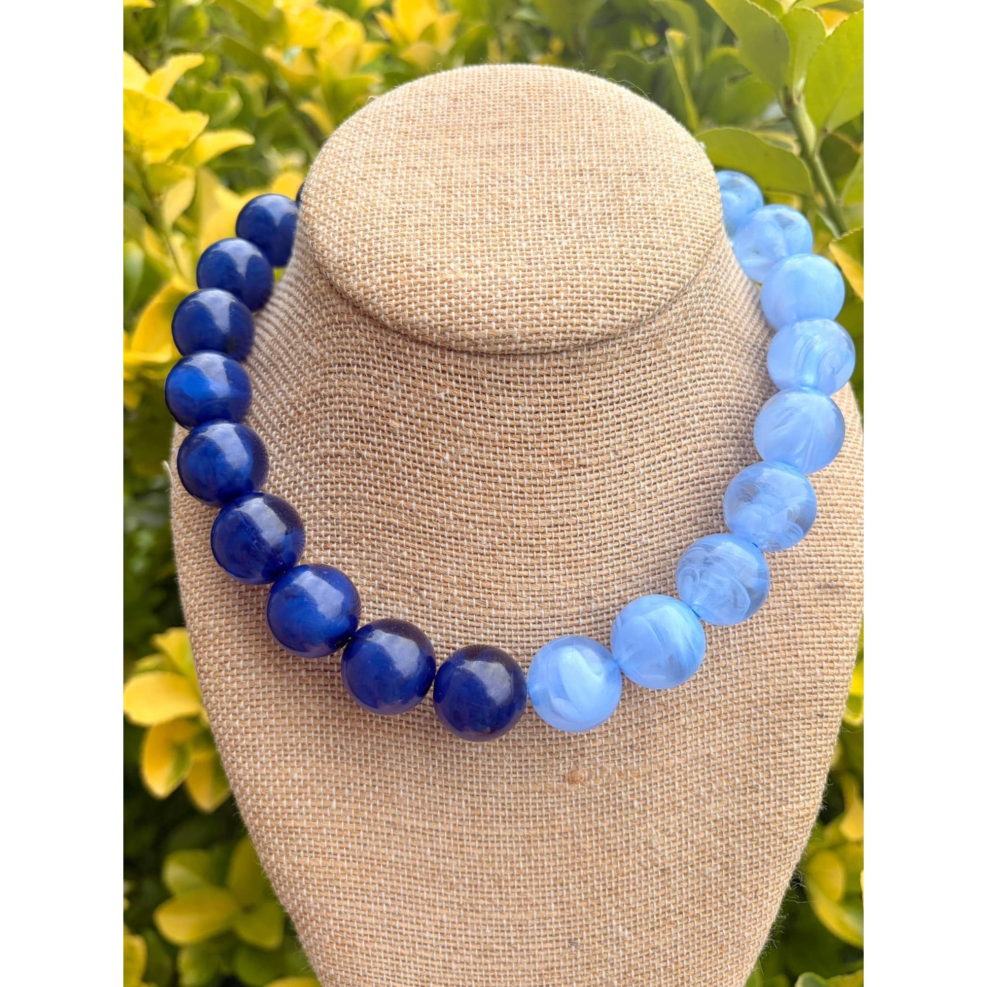 A beaded necklace displayed on a beige fabric jewelry bust, featuring alternating deep cobalt blue and light sky blue round beads, set against a blurred background of yellow flowers and green foliage.