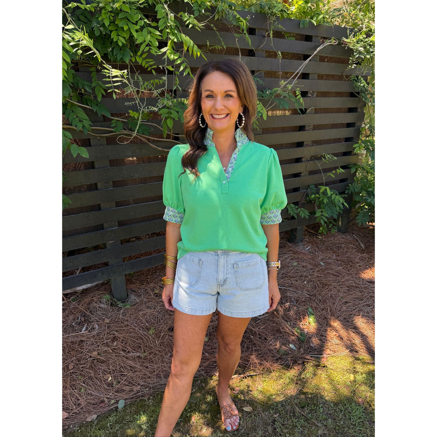 Woman in a green top and denim shorts standing outdoors with plants and a wooden gate in the background.