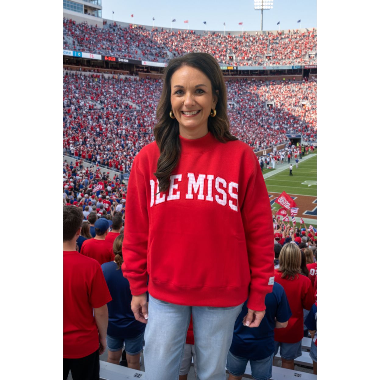 A woman smiling and wearing a bright red crewneck sweatshirt with 'OLE MISS' printed in white arched block letters across the chest, paired with light blue jeans, standing against a plain beige wall.