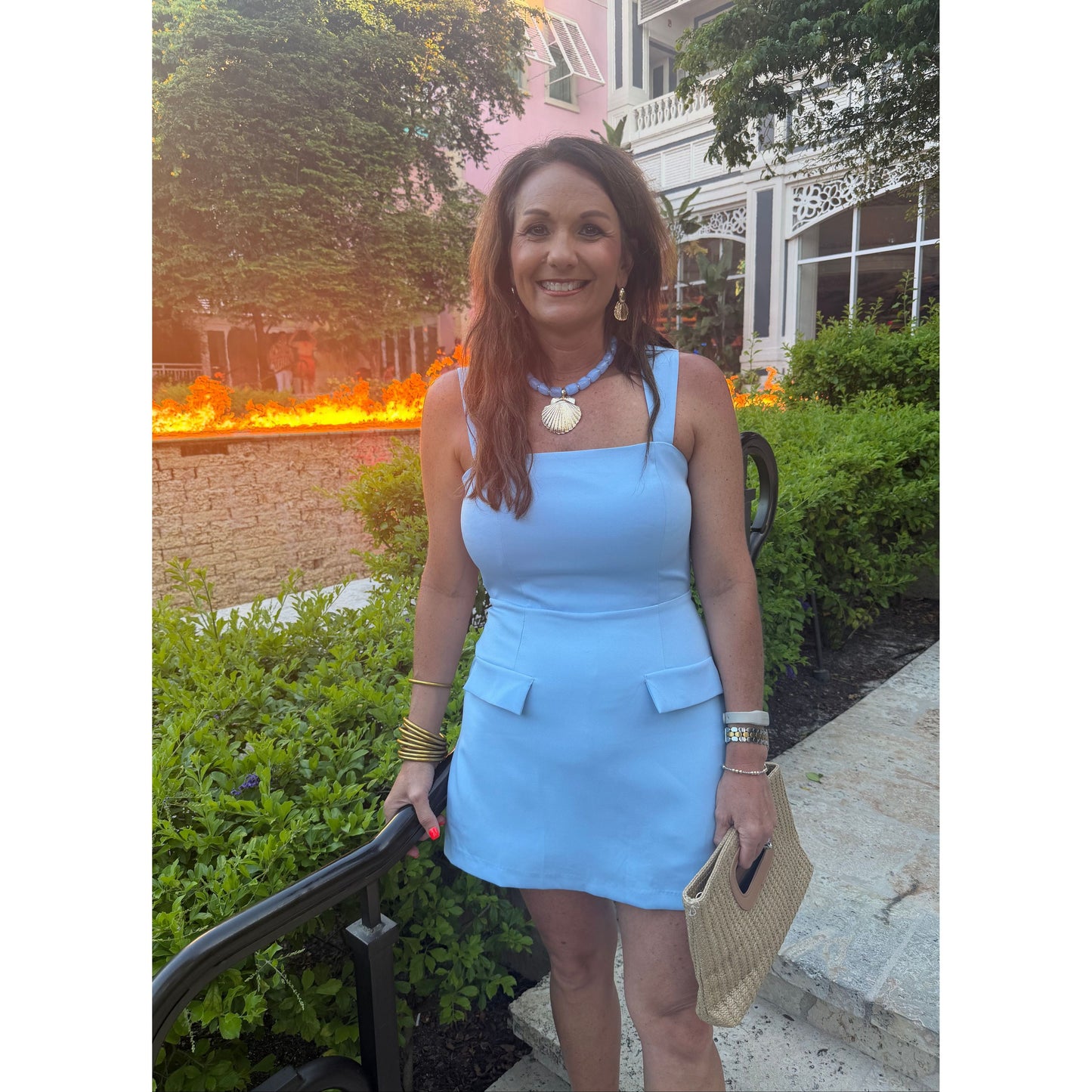 A woman wearing a light blue sleeveless mini dress with shoulder cutouts and flap pockets, standing outdoors on stone steps with greenery and a building in the background.