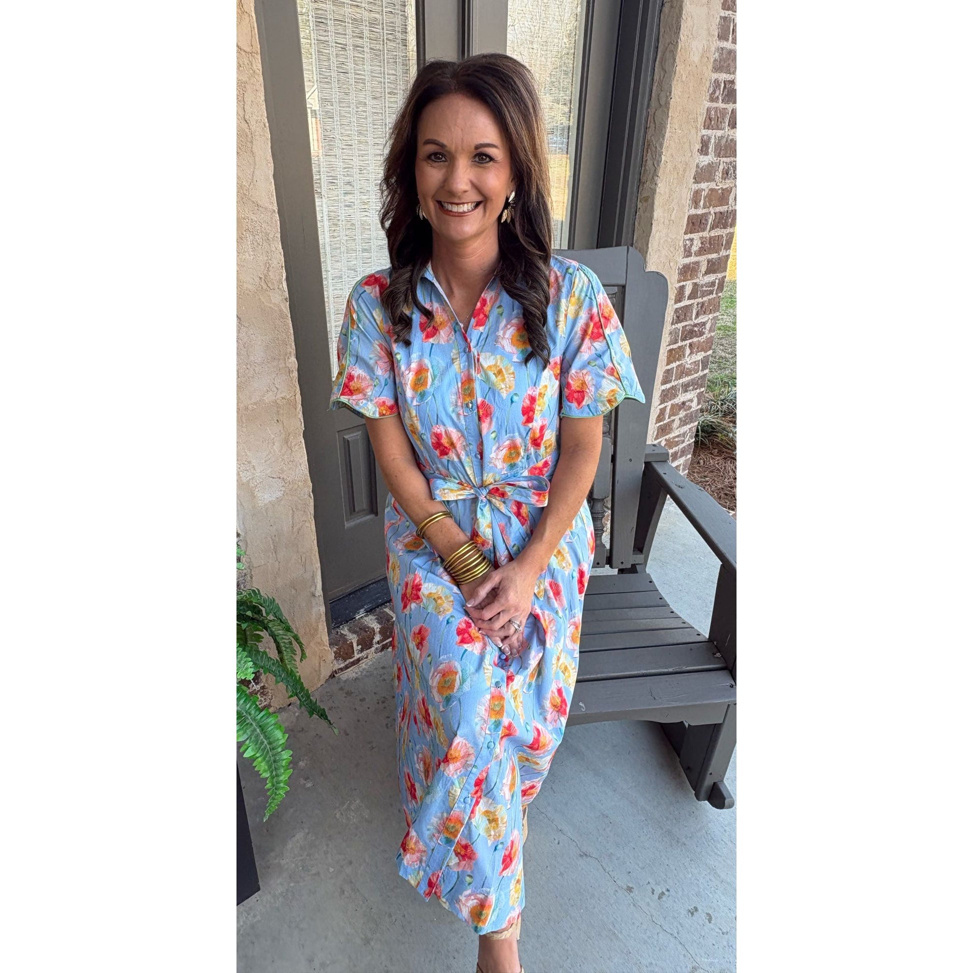 Woman in a floral dress standing on a porch with a plant and bench in the background.