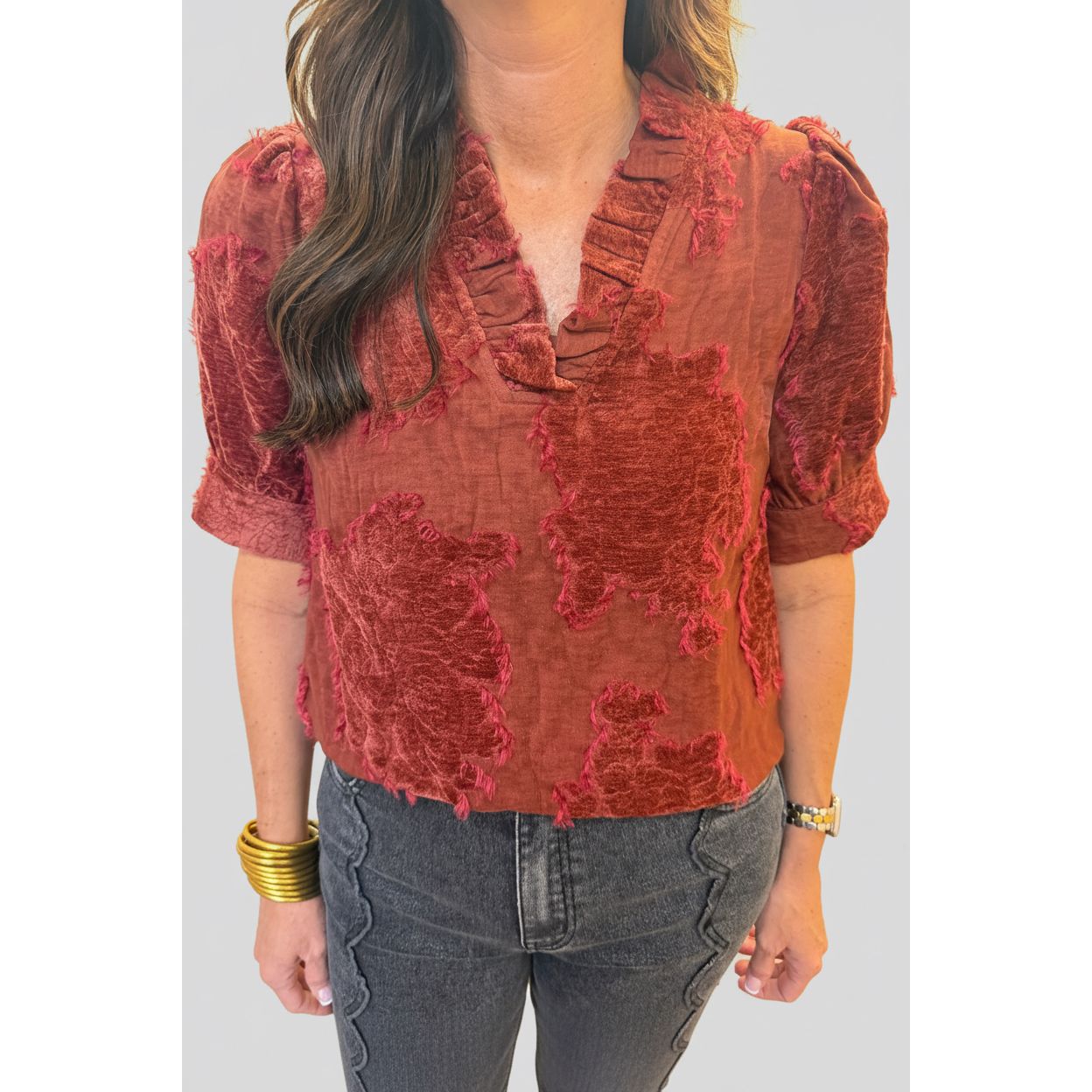 A woman wearing a rust-colored short-sleeve blouse with a textured, distressed pattern and ruffled trim, paired with dark gray jeans.