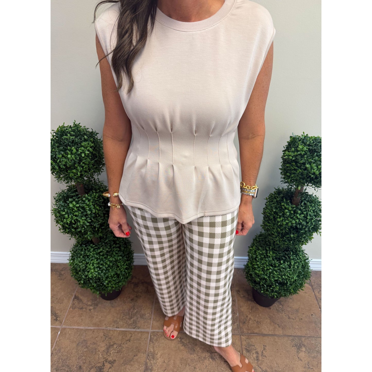 A woman wearing a sleeveless beige knit top with a gathered peplum waist and brown-and-white gingham cropped pants, standing indoors between two green topiary trees.