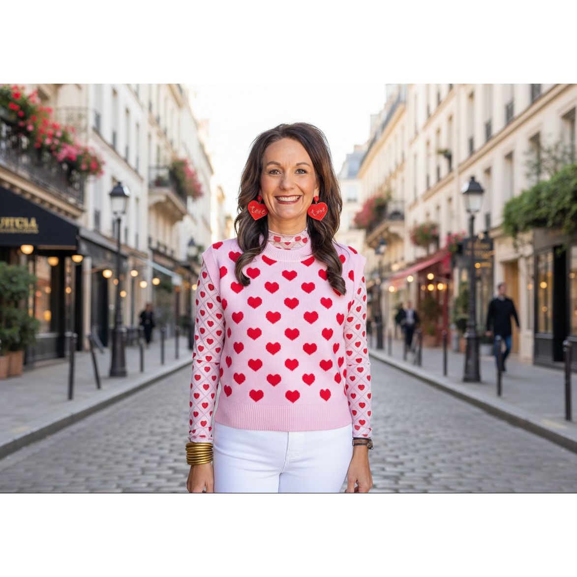 Woman wearing a pink sweater with red heart patterns and heart-shaped earrings, standing in on a street.