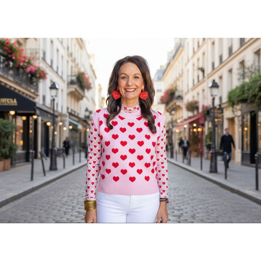 Woman wearing a pink sweater with red heart patterns and heart-shaped earrings, standing in on a street.