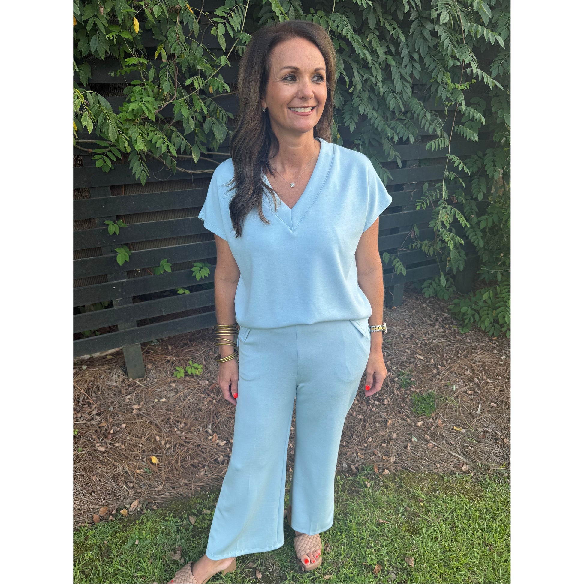 A woman wearing a light blue short-sleeve V-neck top and matching wide-leg pants, standing outdoors on a bench with greenery and mulch in the background.