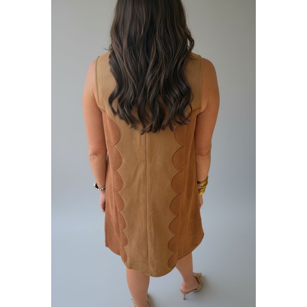 A woman viewed from behind wearing a sleeveless tan dress with scalloped cutout details along the sides, standing on a covered walkway.