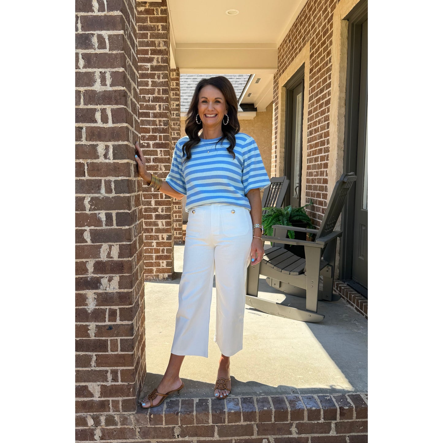 Woman in a blue striped shirt and white pants standing on a brick porch.