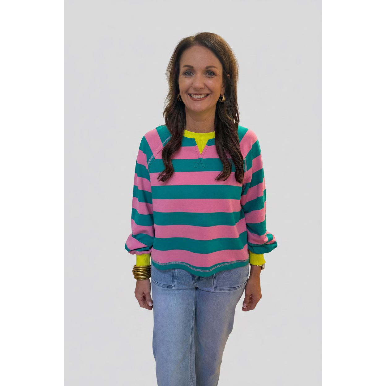 A woman stands indoors wearing a long-sleeve knit top with bold horizontal pink and teal stripes, a bright yellow-green V-neck trim, and matching yellow-green cuffs, paired with light blue jeans.