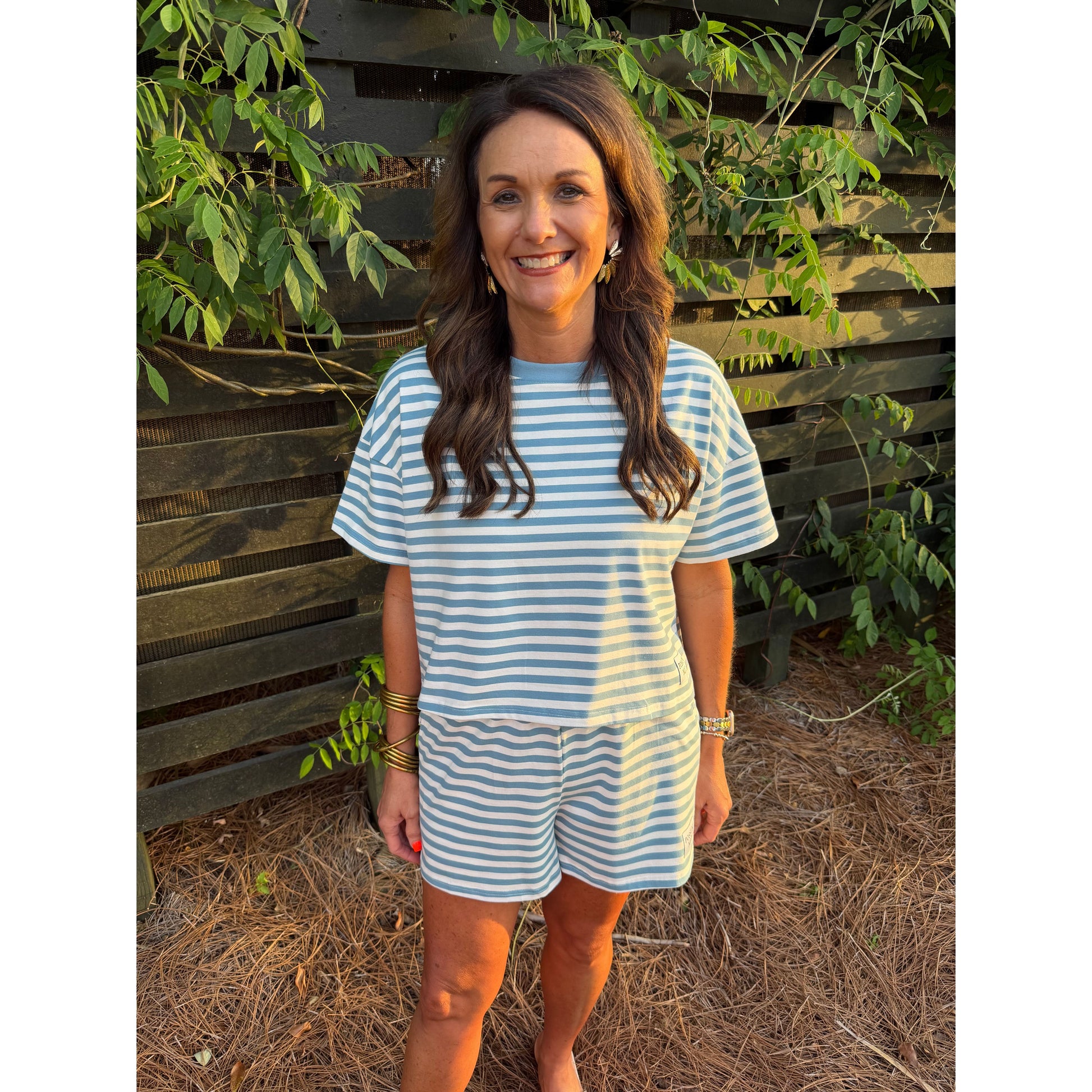 Woman wearing a blue and white striped outfit standing in front of a wooden fence with greenery.