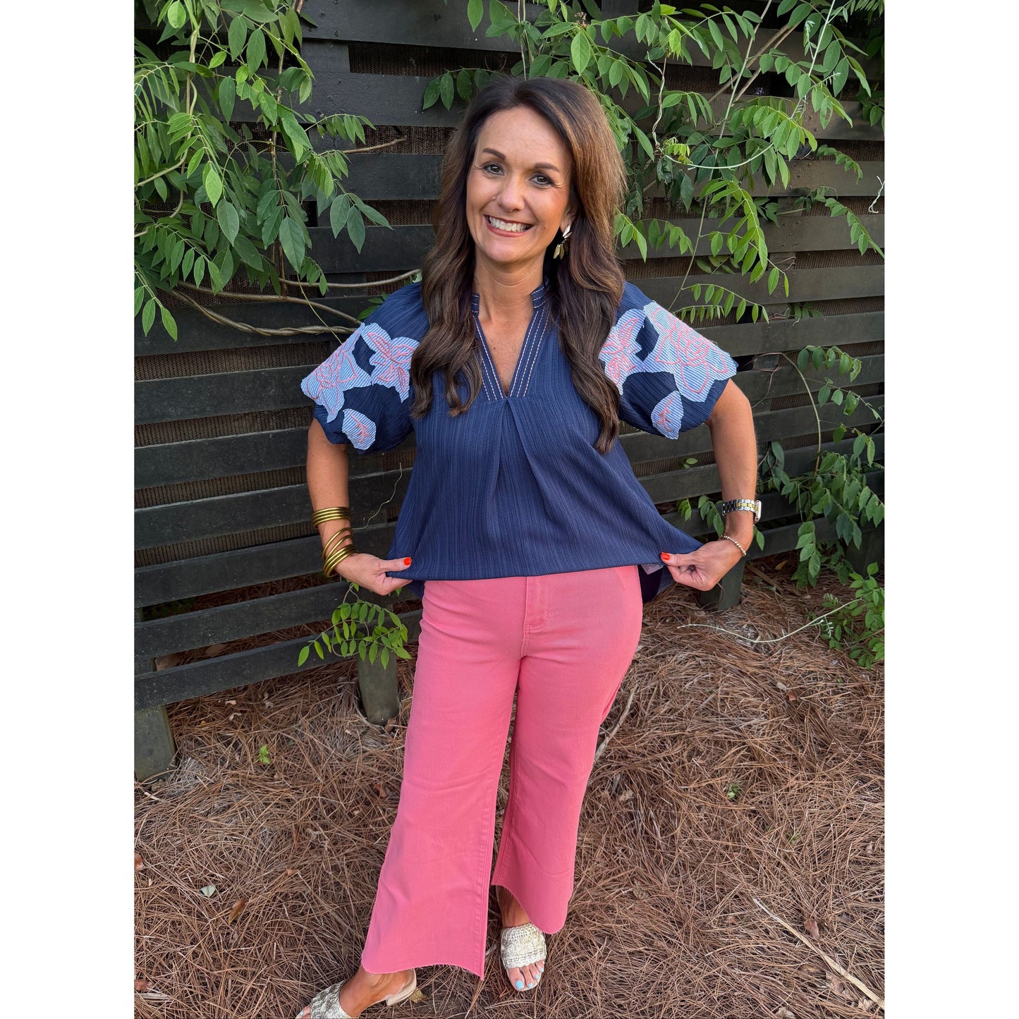 Woman wearing a blue top with floral sleeves and pink pants standing in front of a wooden fence with greenery.