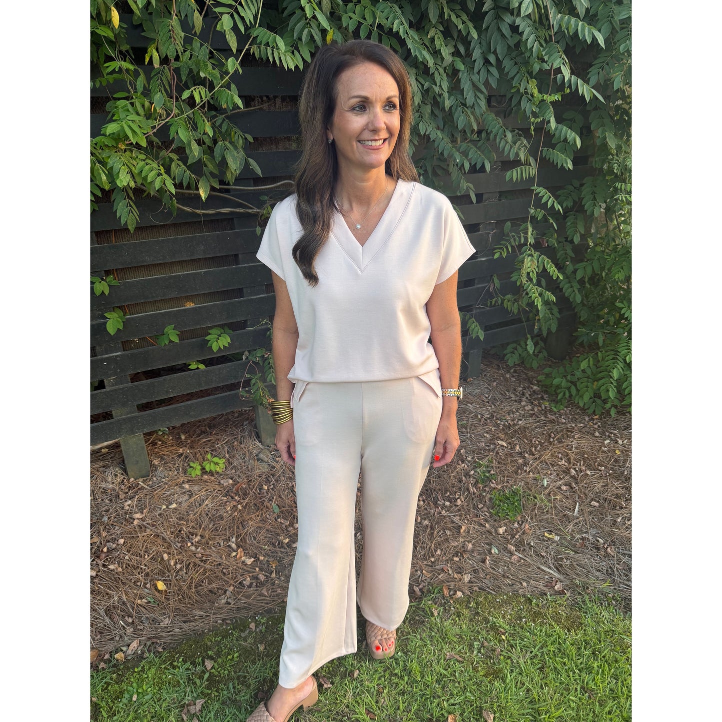 A woman wearing a cream-colored short-sleeve V-neck top and matching wide-leg pants, standing outdoors on grass in front of a dark wooden fence and green foliage.