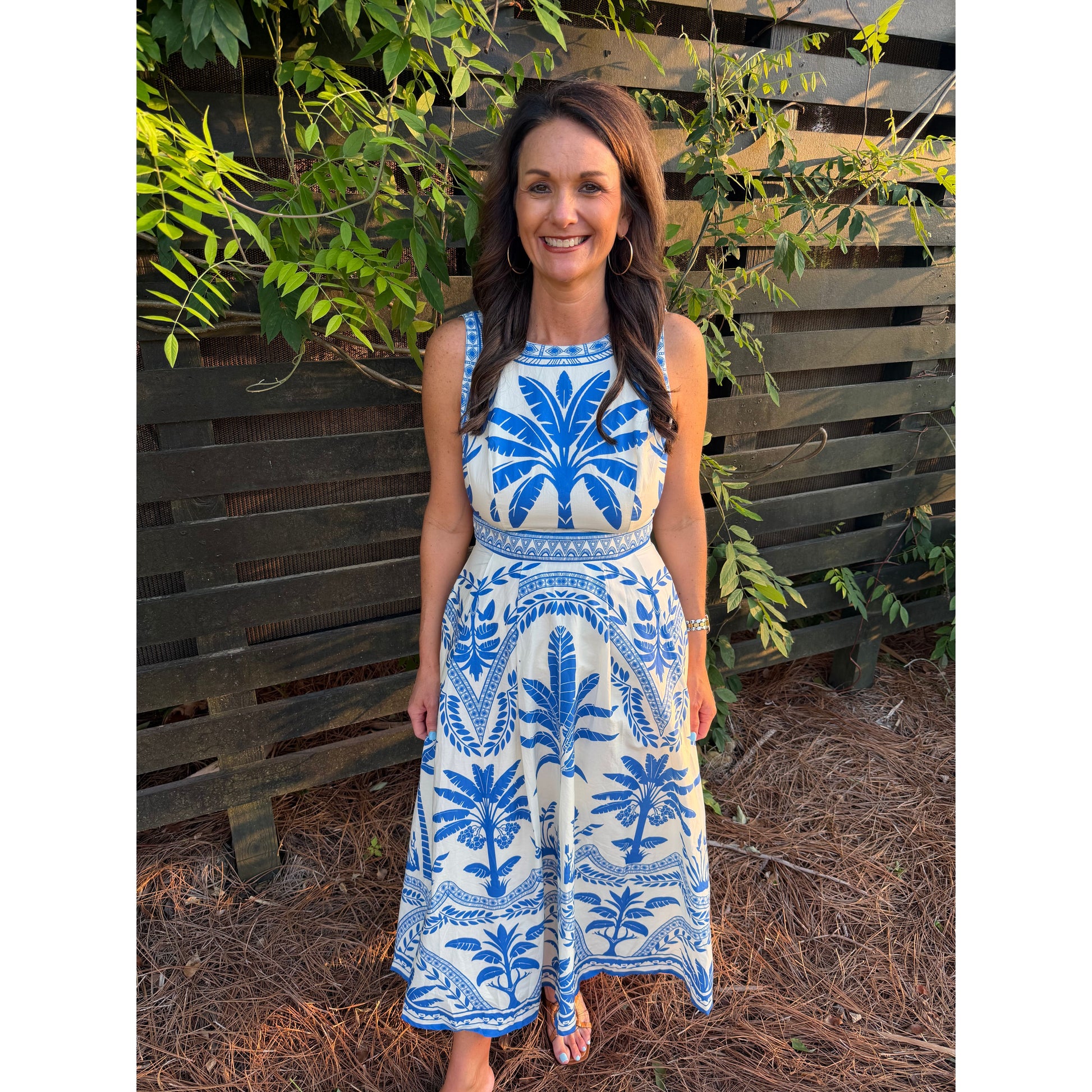 Woman wearing a blue and white patterned dress standing in front of a wooden fence with greenery.