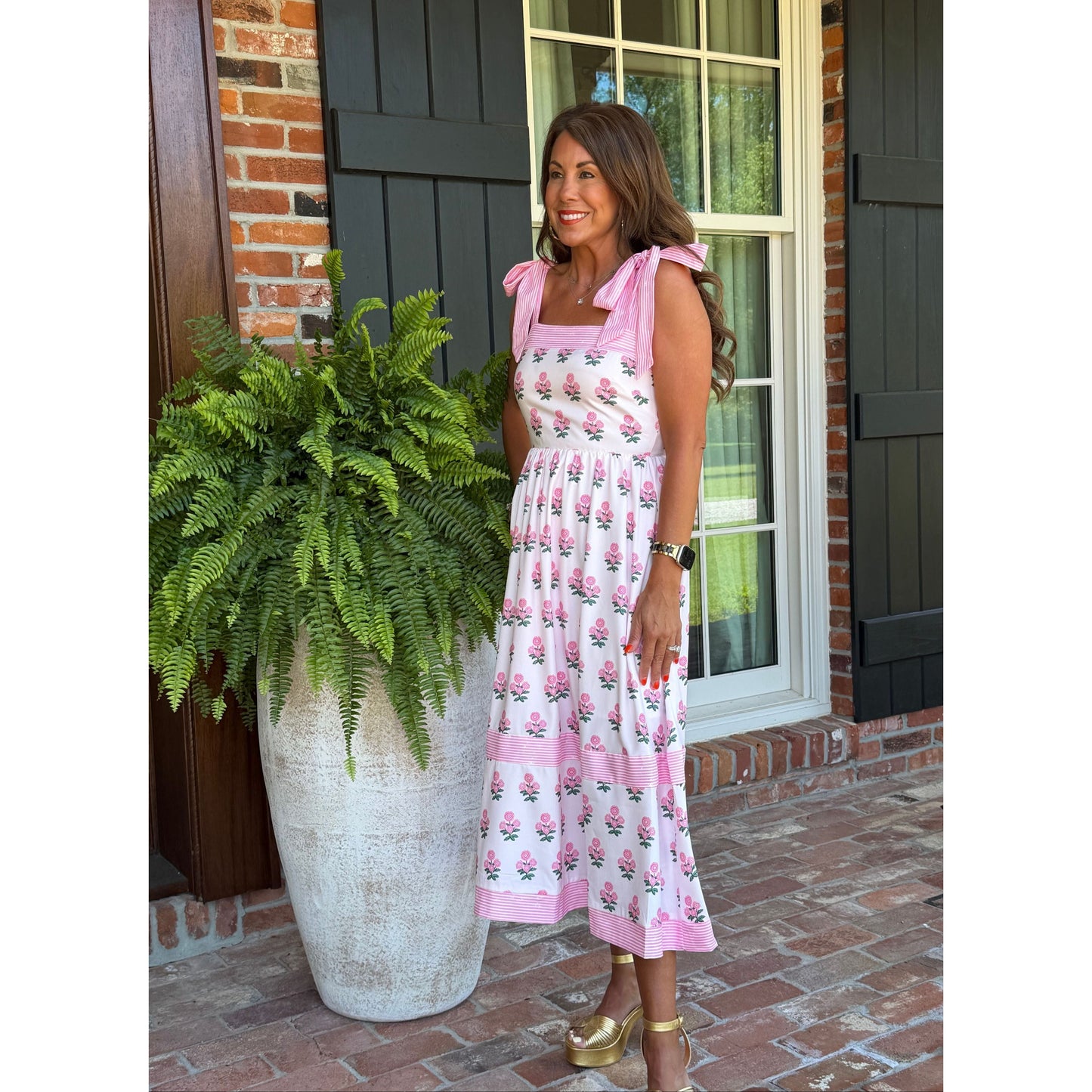 Woman in a pink floral dress standing next to a large planter with green ferns on a porch.