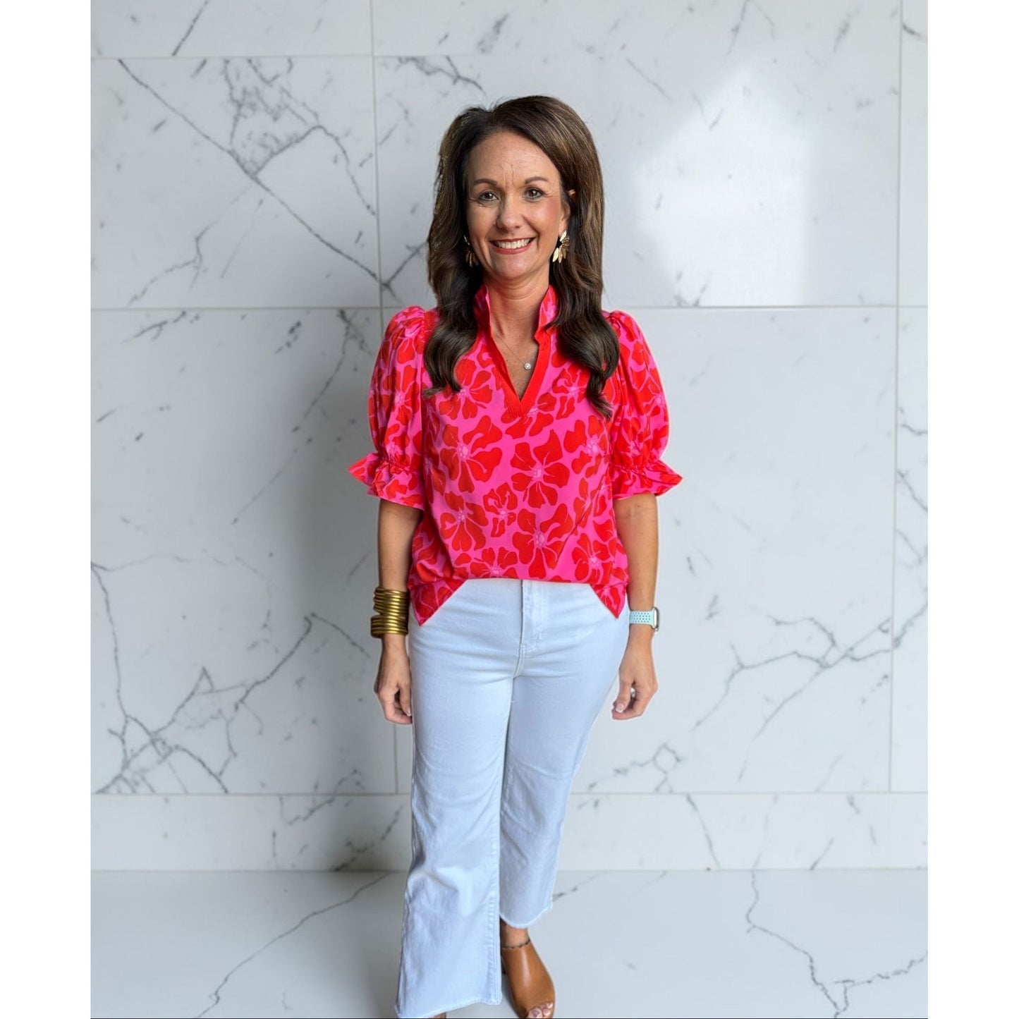 Woman wearing a bright pink blouse with ruffled sleeves and white pants against a marble wall.
