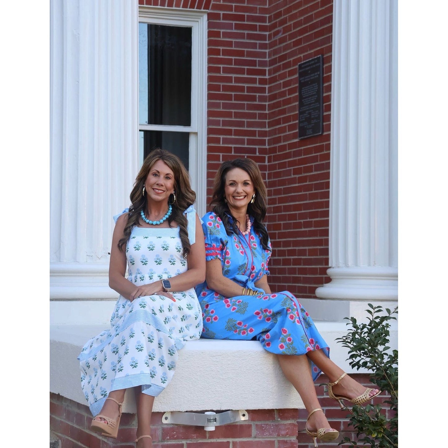 Two women sitting on a bench in front of a brick building with white columns.