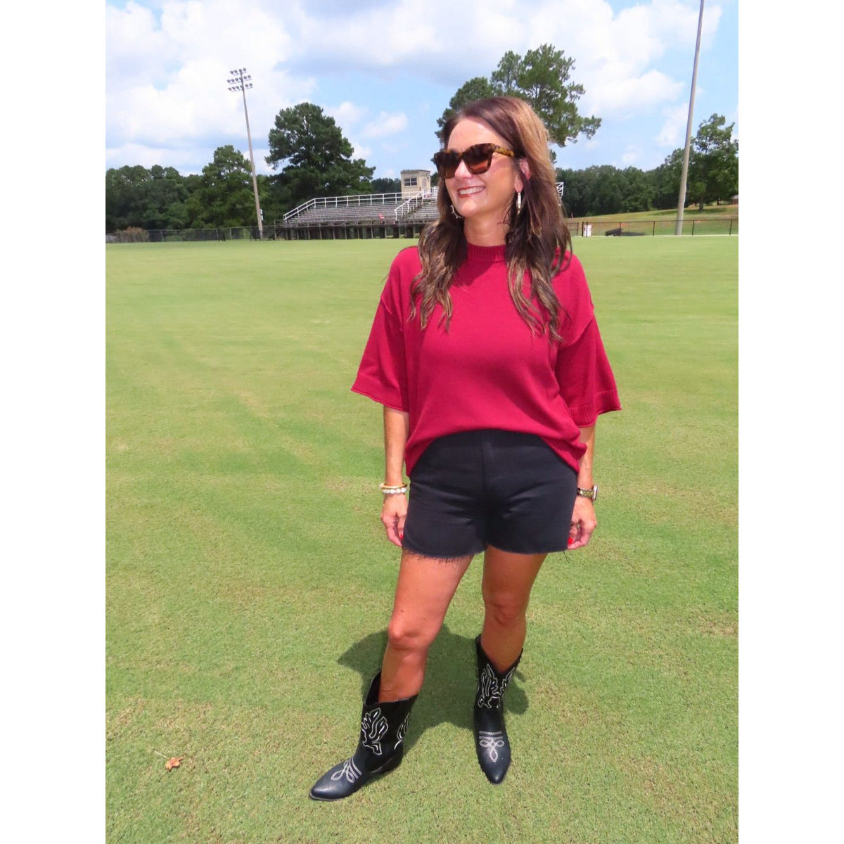 A woman stands on a grassy field wearing a bright red oversized short-sleeve top, black shorts, and black western-style boots with white embroidery. She is also wearing sunglasses and jewelry.