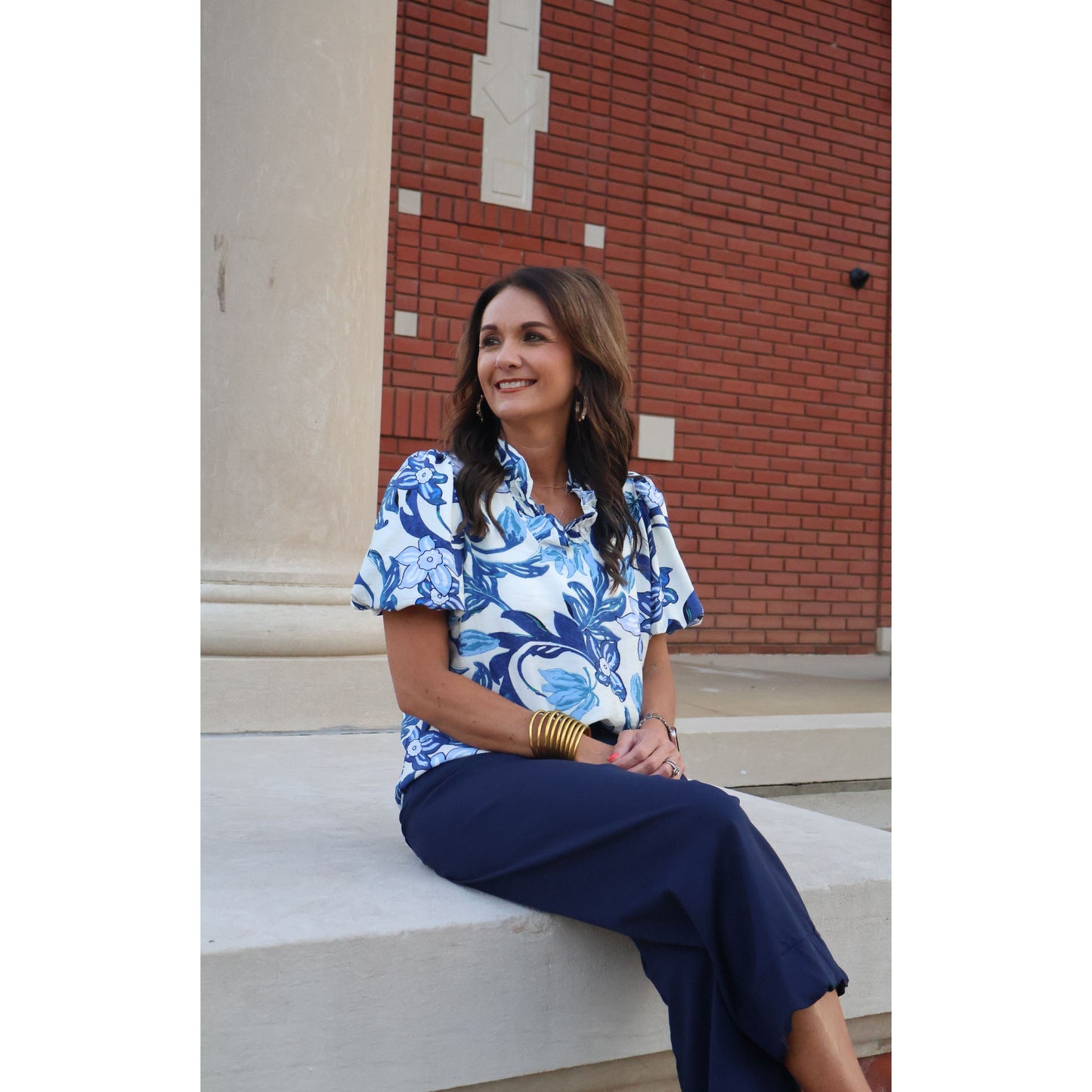 Woman sitting on steps wearing a blue and white patterned blouse and navy pants, with a brick building in the background.