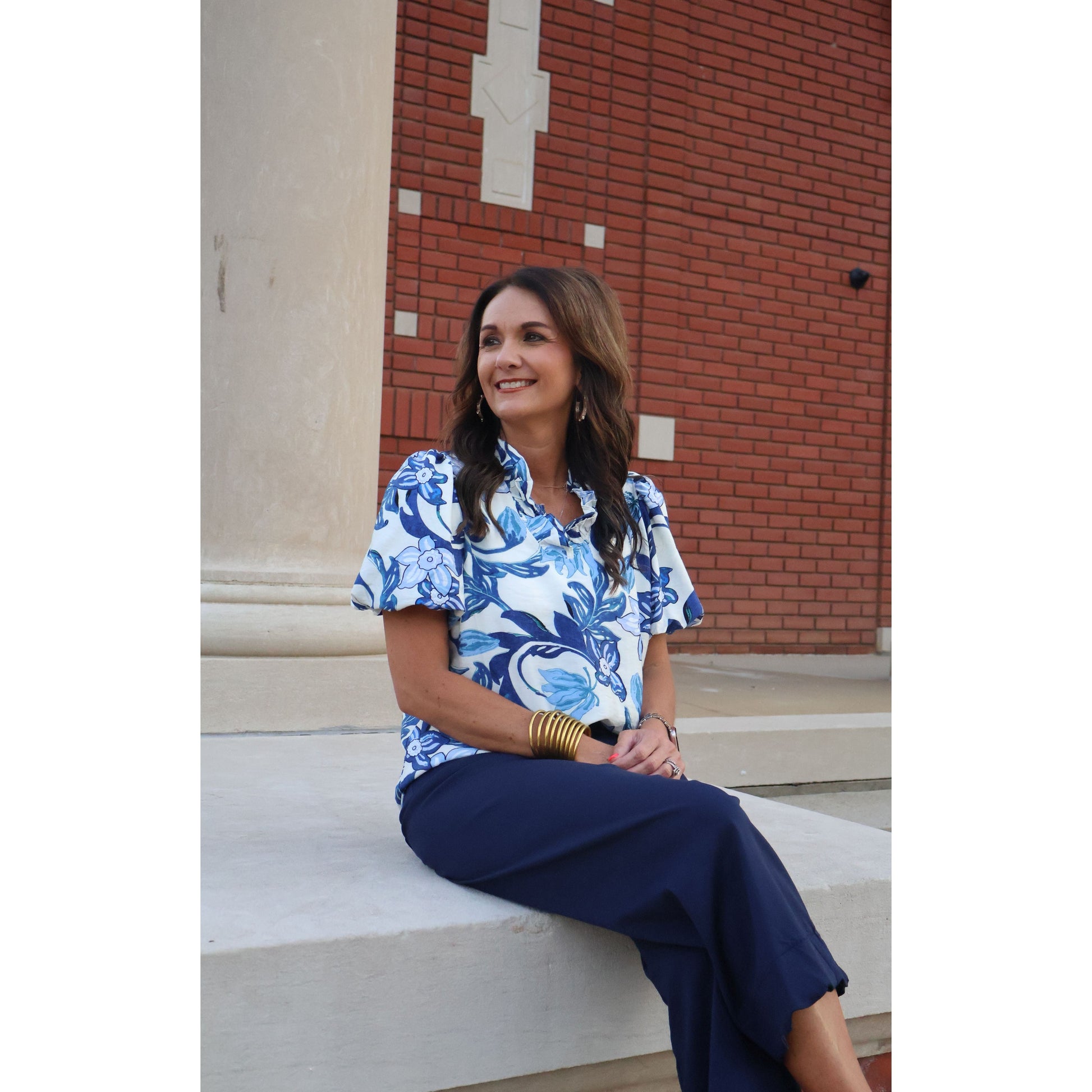 Woman sitting on steps wearing a blue and white patterned blouse and navy pants, with a brick building in the background.