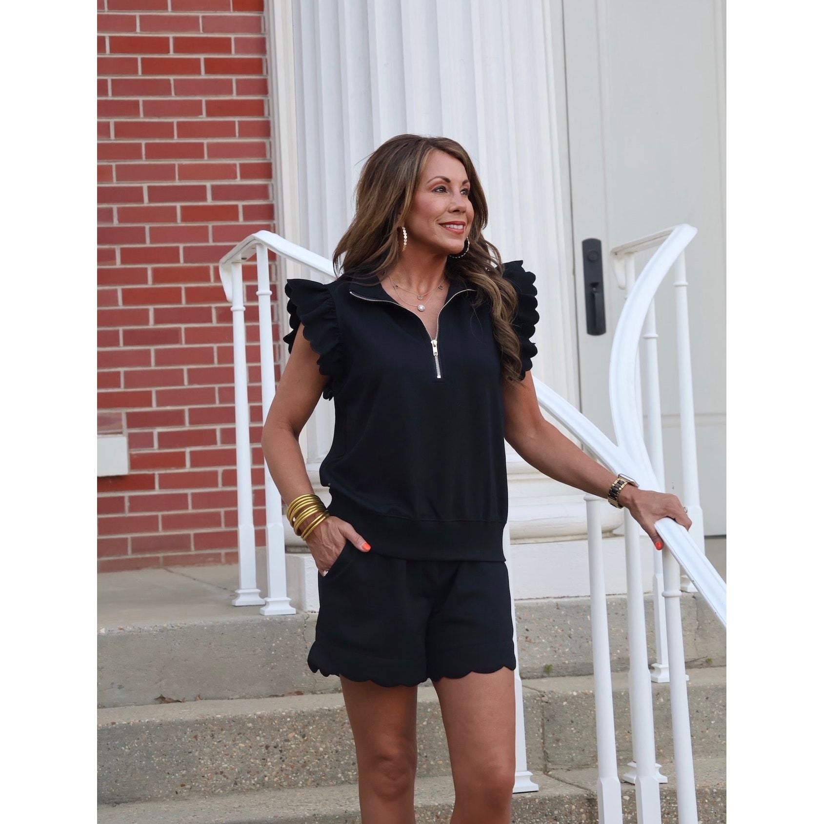 Woman in a black outfit standing on steps with a white railing and brick wall background