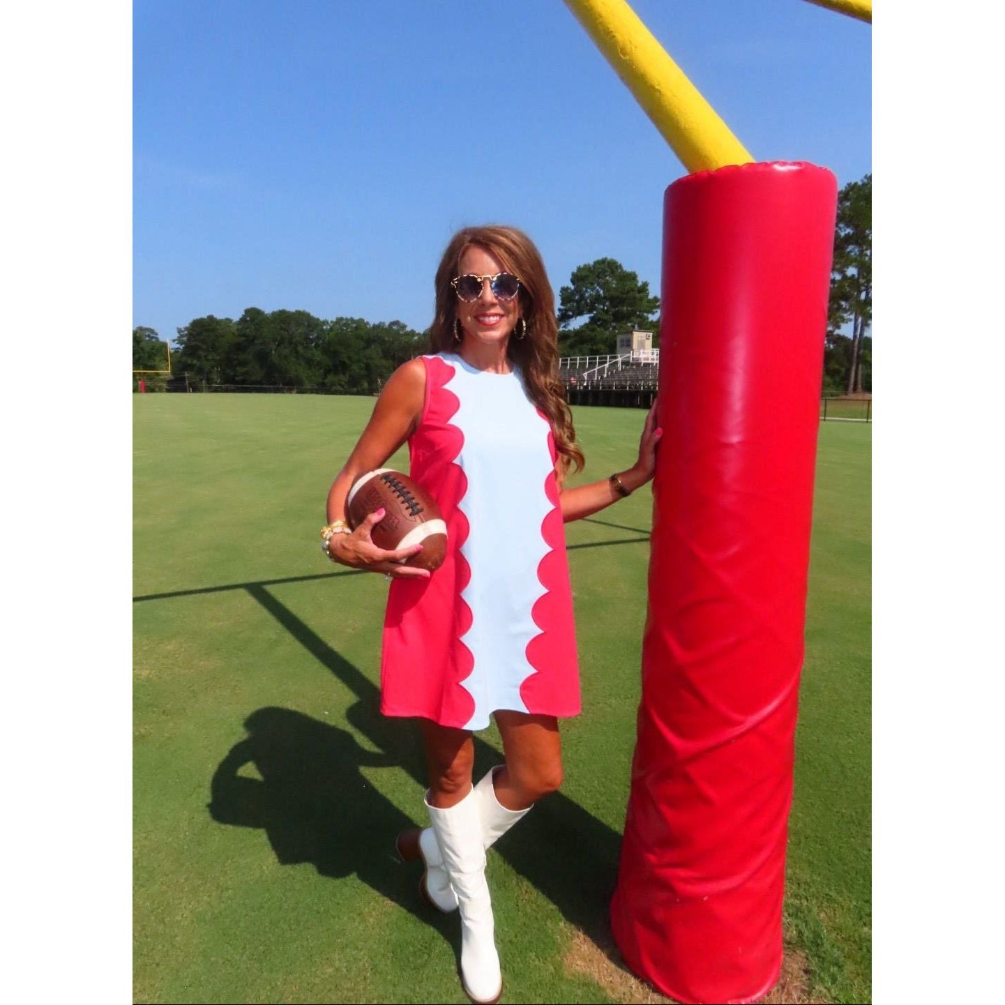 A woman stands on a grassy field wearing a sleeveless red and white dress with scalloped edges, white knee-high boots, and sunglasses, holding a football next to a large red goal post.