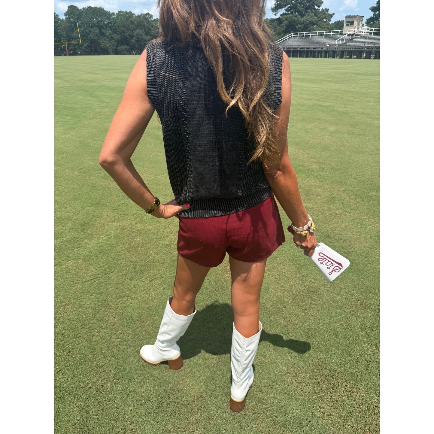 A woman stands on a grassy field wearing a sleeveless black ribbed knit top, burgundy shorts, and white cowboy boots, holding a white phone case.