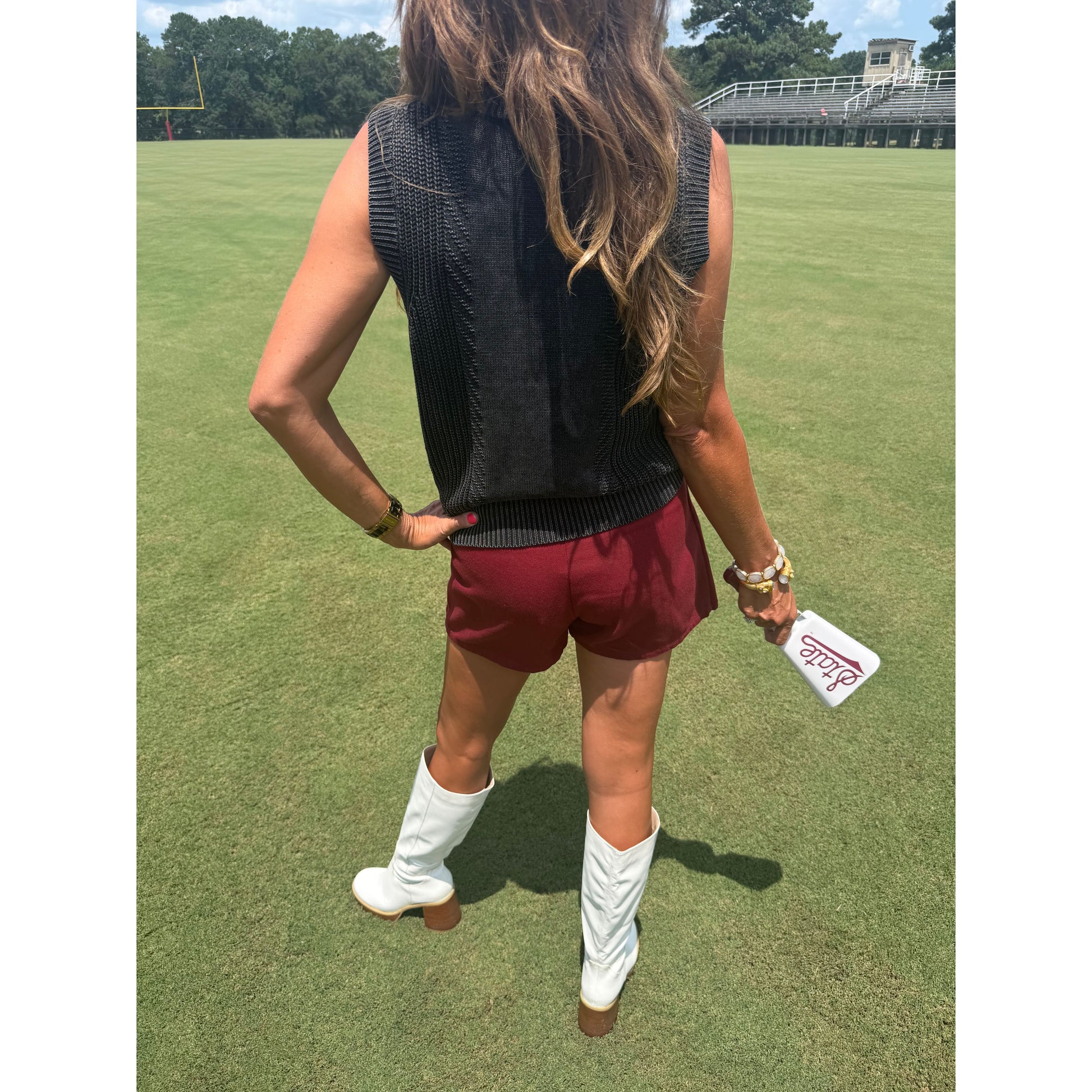 A woman stands on a grassy field wearing a sleeveless black ribbed knit top, burgundy shorts, and white cowboy boots, holding a white phone case.