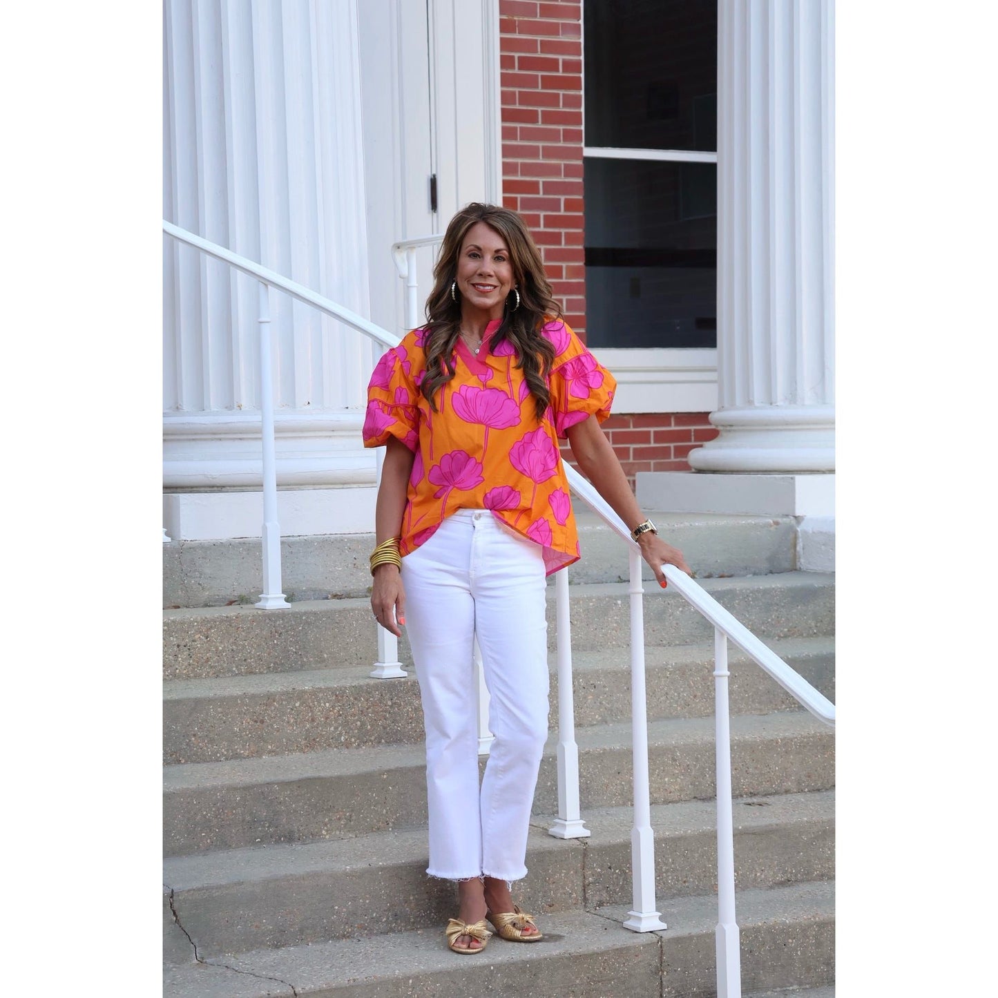 Woman in a bright orange blouse with pink floral patterns and white pants standing on steps.
