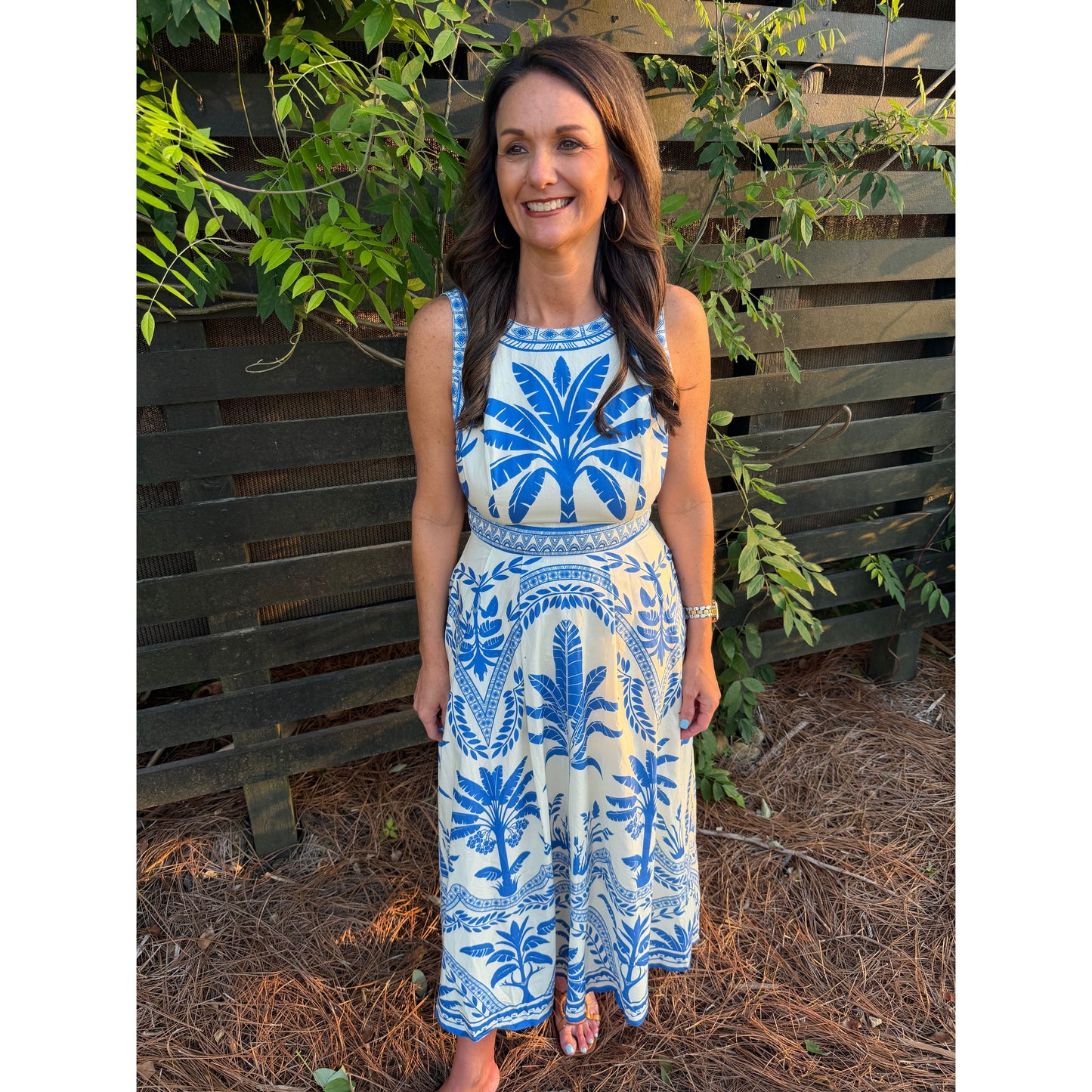 Woman wearing a blue and white patterned dress standing outdoors with greenery in the background