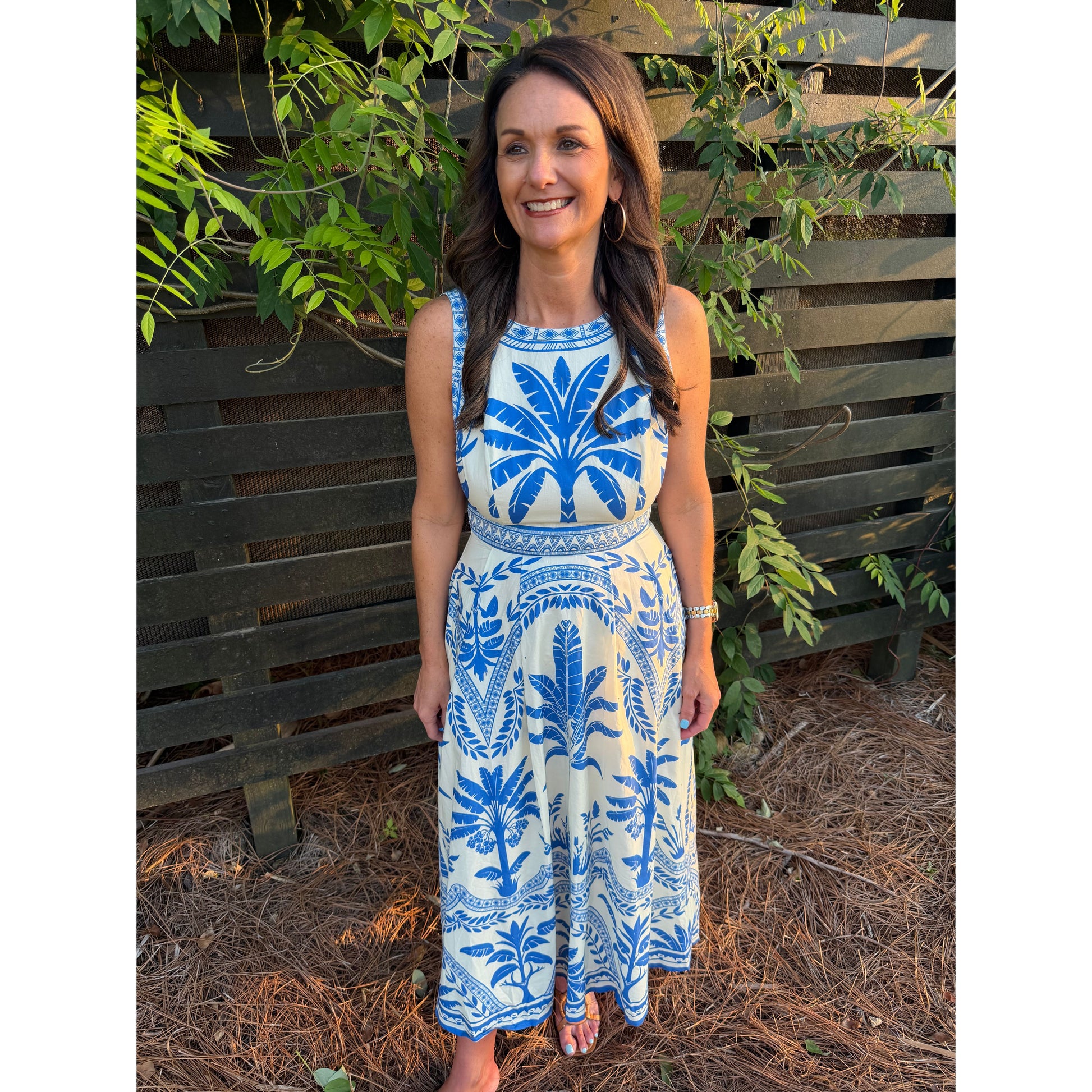 Woman wearing a blue and white patterned dress standing outdoors with greenery in the background