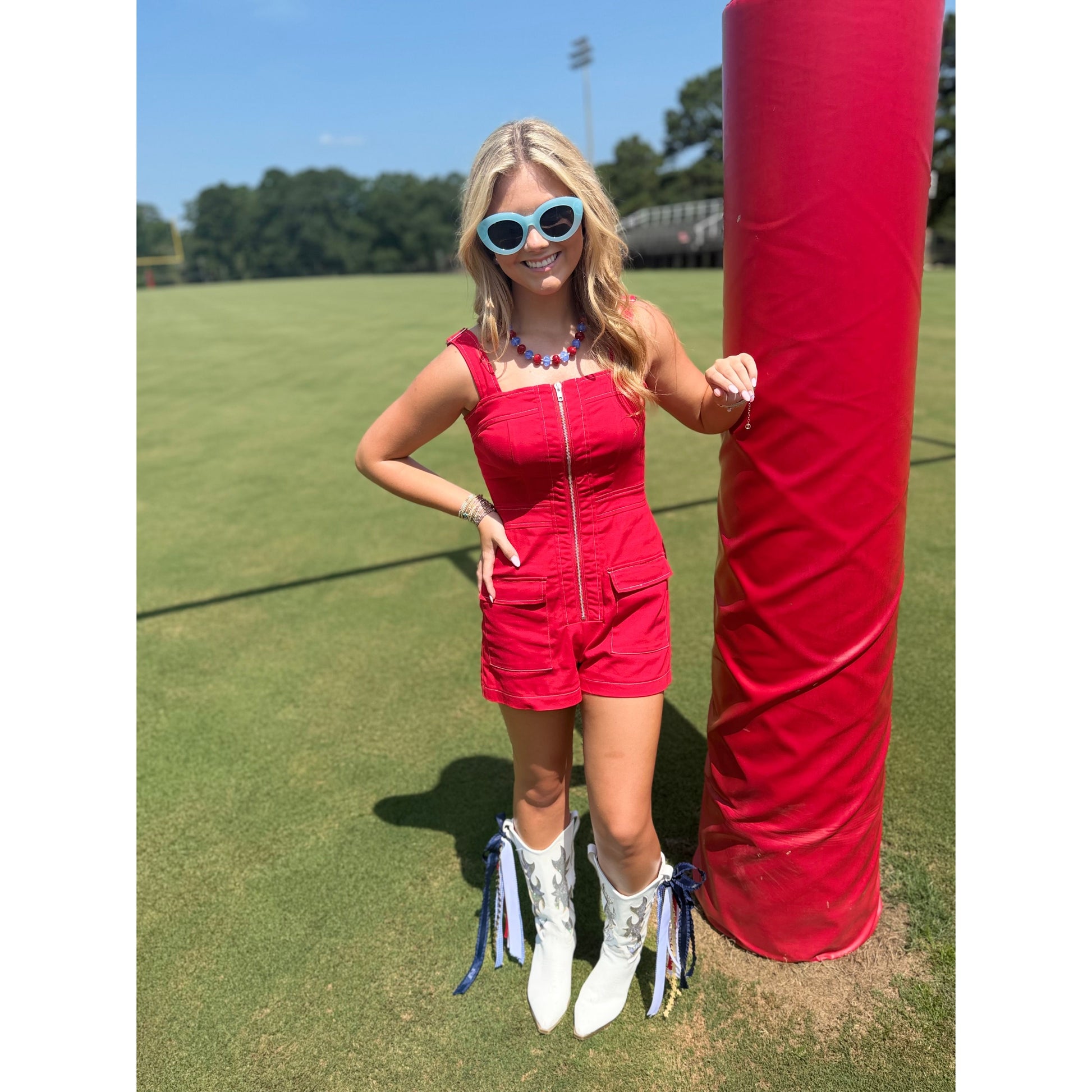 A woman stands outdoors on a grassy field next to a large red padded pole, wearing a bright red sleeveless romper with a front zipper, white western-style boots with blue tassels, and light blue sunglasses.