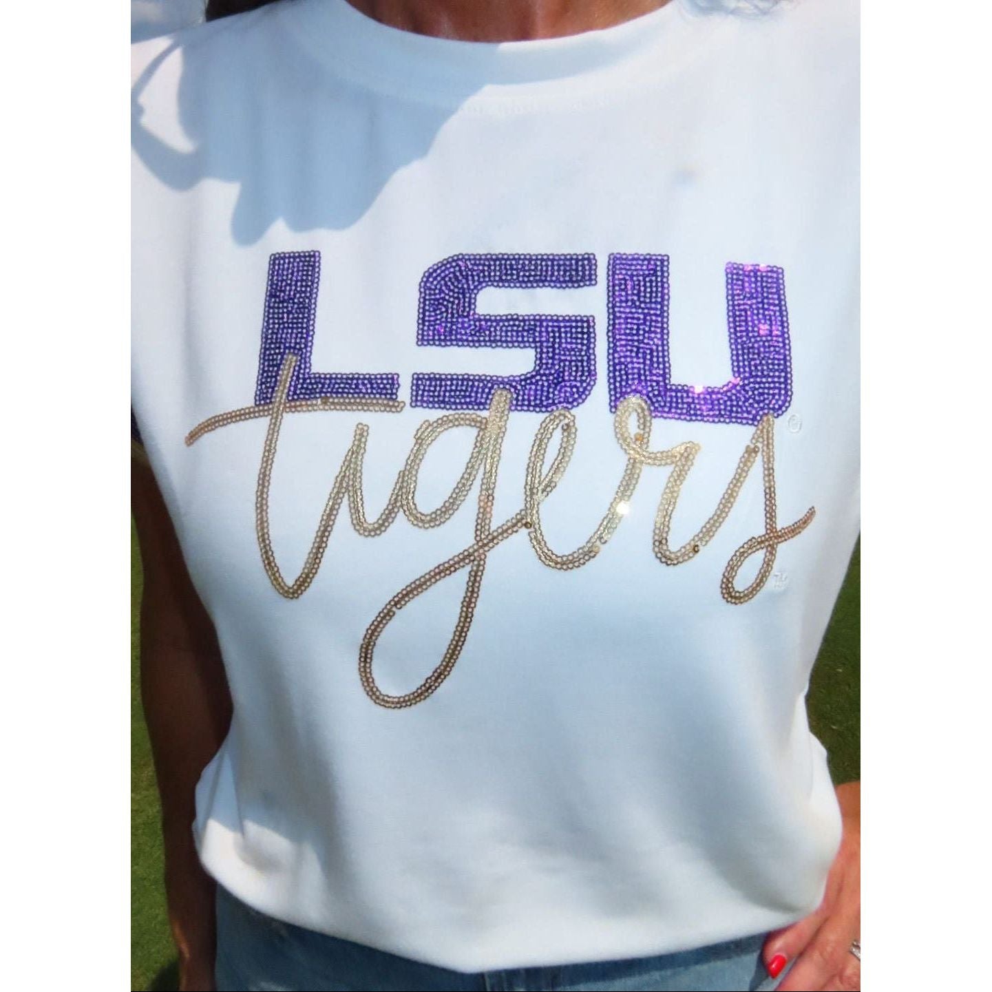 A white short-sleeve t-shirt featuring a rhinestone-embellished graphic that reads 'LSU' in purple block letters and 'tigers' in gold cursive script, worn by a model outdoors.