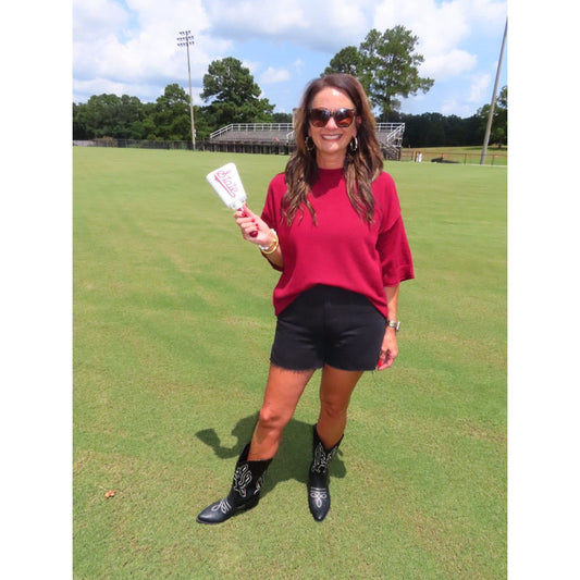 A woman stands on a grassy field holding a white and red golf club, wearing a red t-shirt, black shorts, and black western boots.