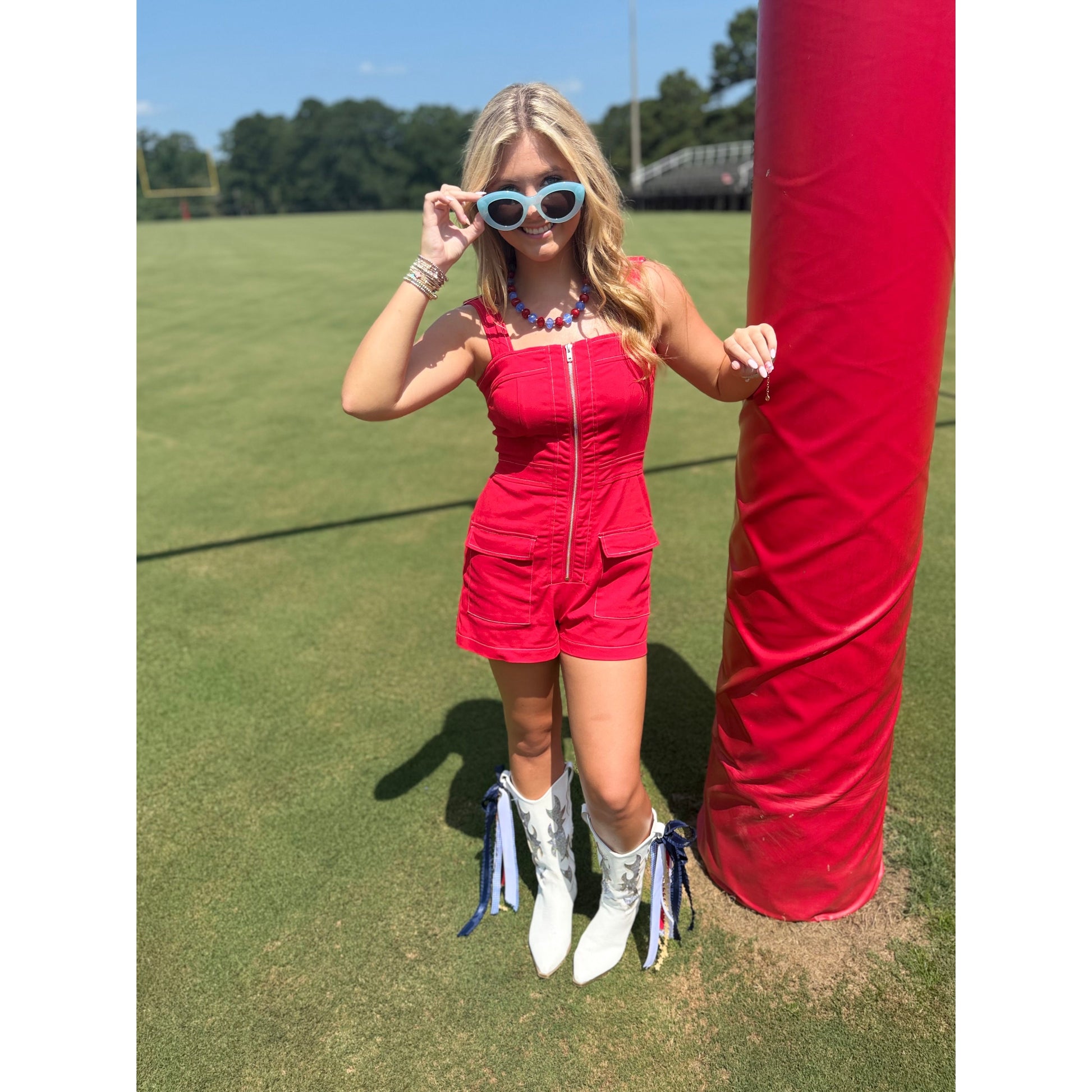 A woman stands on a grassy field wearing a bright red sleeveless romper with a front zipper, white cowboy boots with blue and multicolor tassels, and light blue sunglasses. She is posing next to a large red padded post.