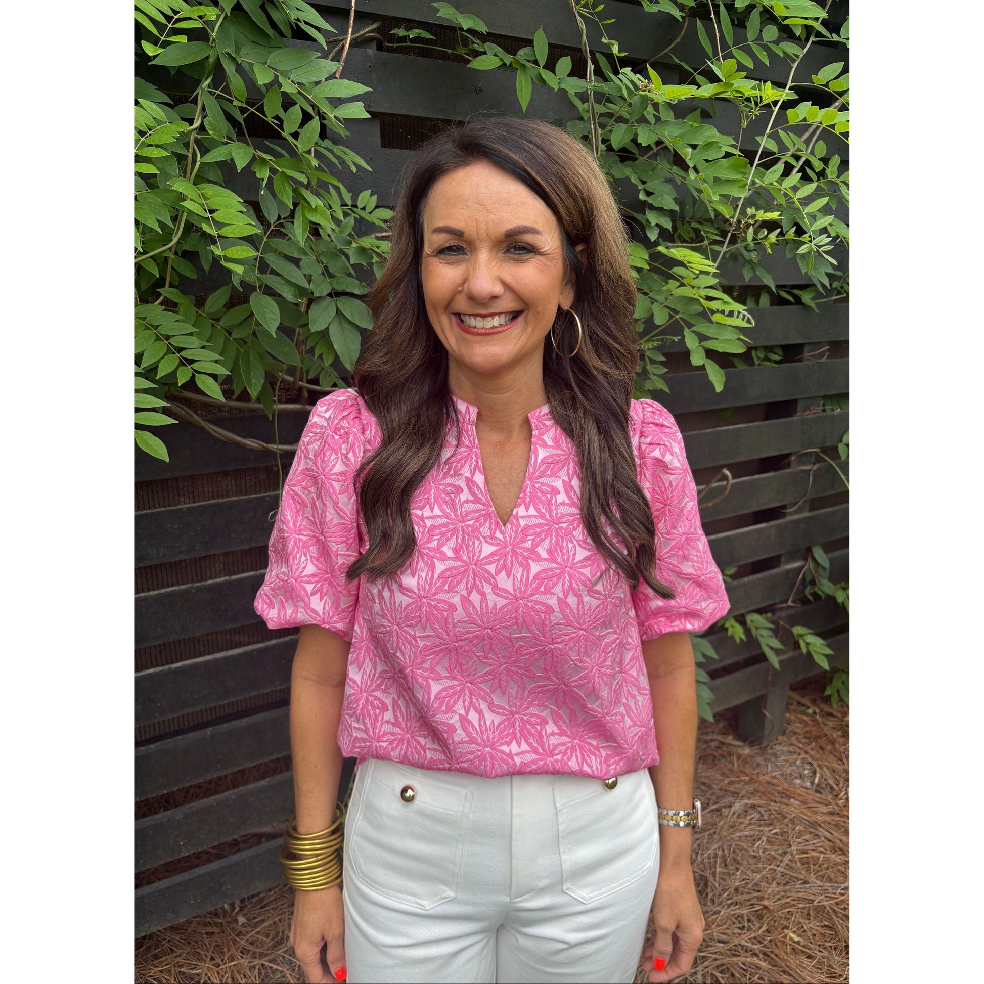 Woman wearing a pink blouse with a floral pattern, sitting outdoors with plants around.